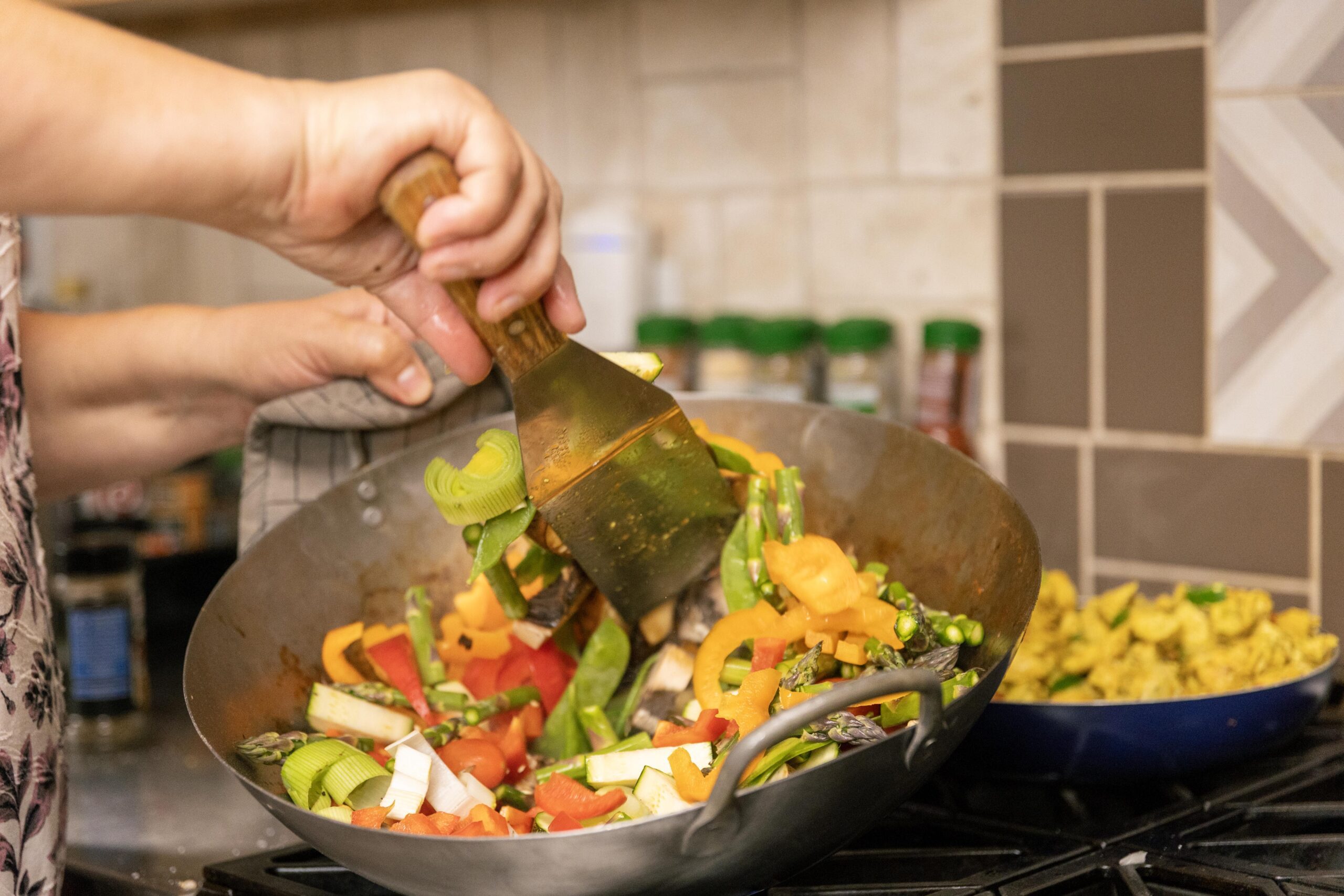 Person cooking a colorful mix of fresh vegetables, including bell peppers, asparagus, and zucchini, in a wok on a stovetop, with a bowl of seasoned food in the background.