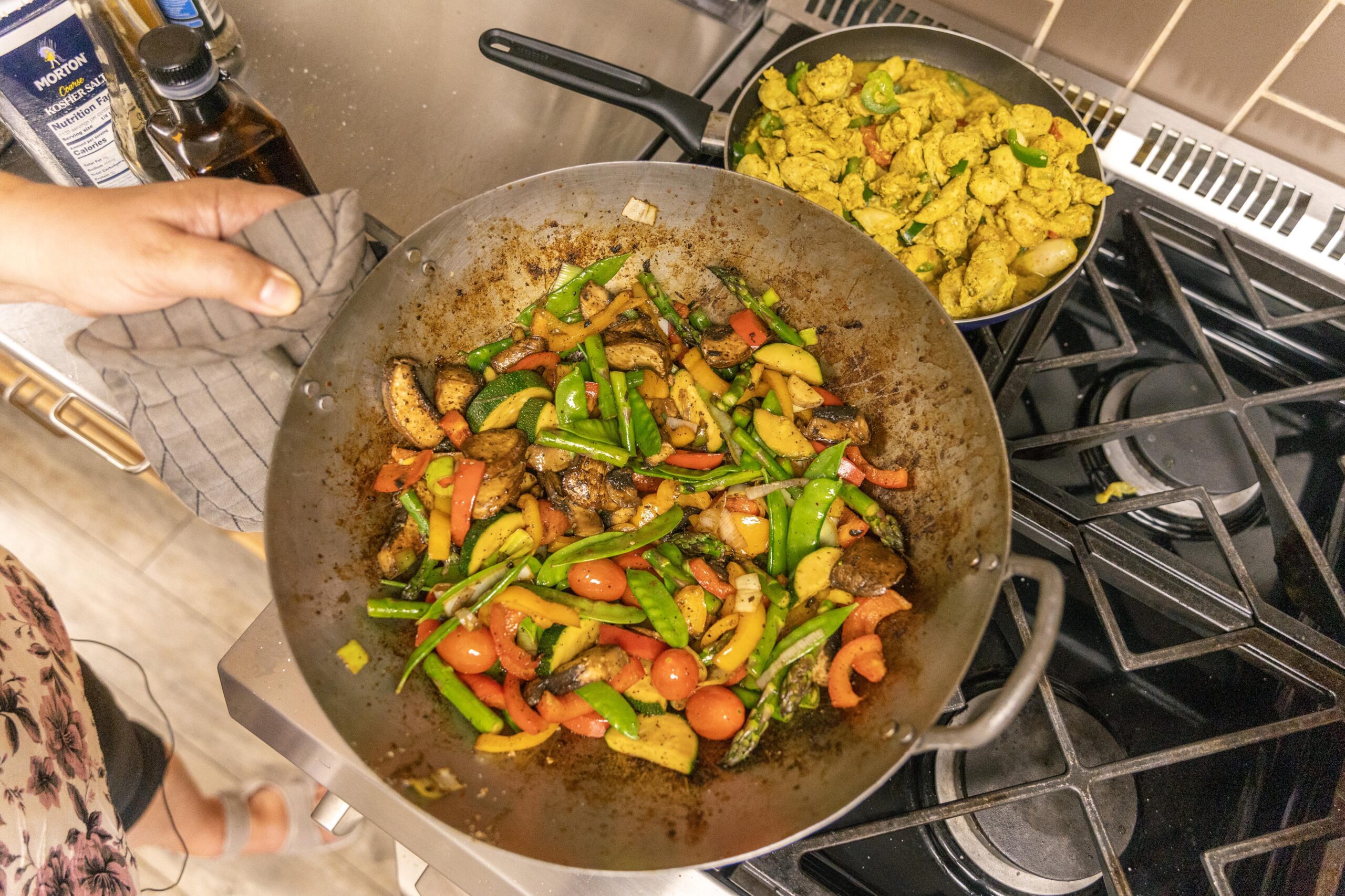Colorful stir-fried vegetables including bell peppers, green beans, and cherry tomatoes in a wok, with a separate pan of seasoned chicken in the background, showcasing a vibrant cooking scene.
