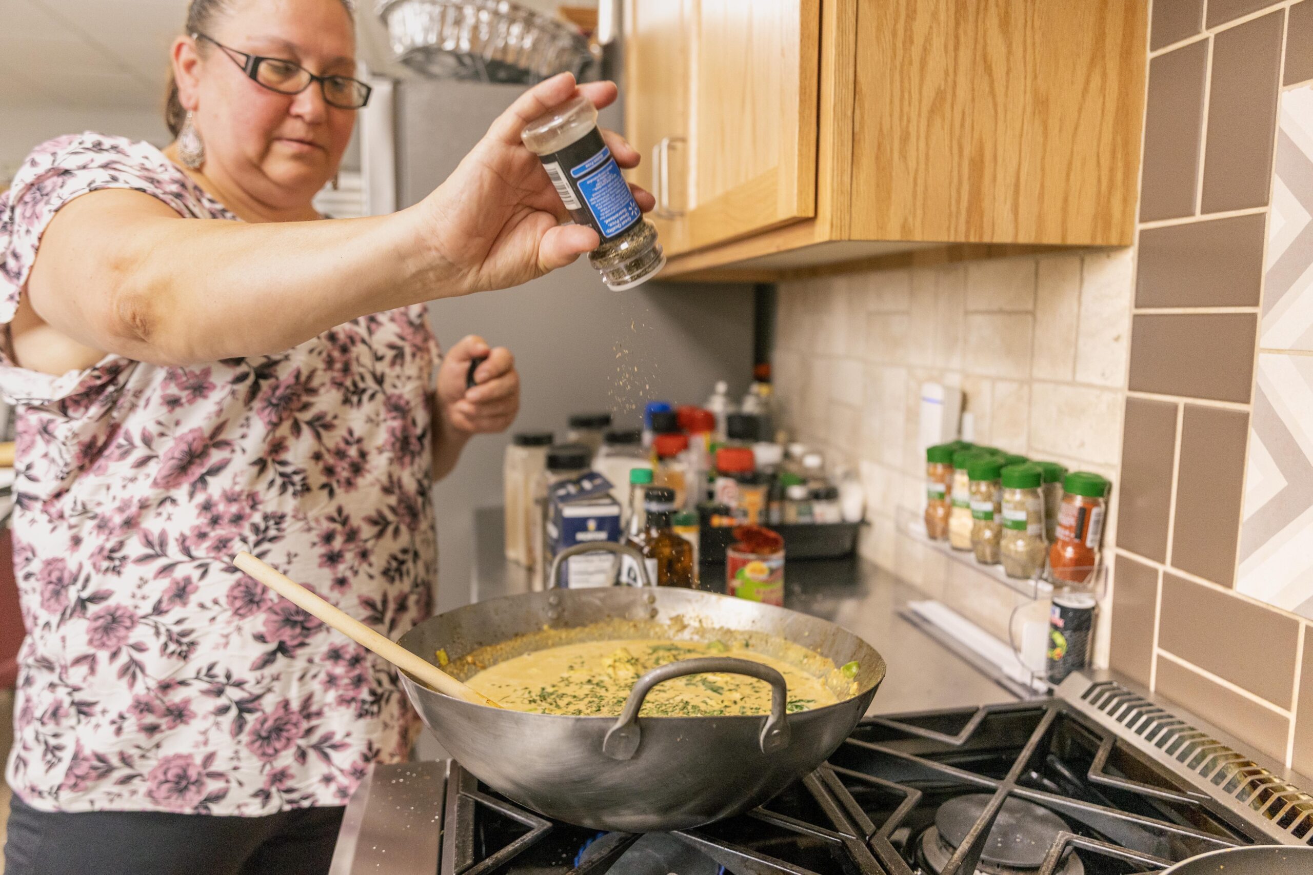 Woman adding spices to a large pot of soup on the stovetop, surrounded by various cooking ingredients and spices in a modern kitchen.