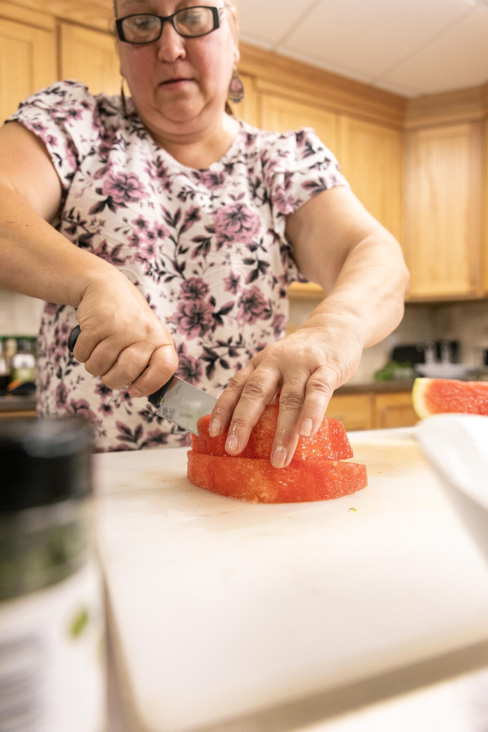Woman slicing watermelon in a kitchen with wooden cabinets, showcasing fresh fruit preparation for a healthy meal or snack.