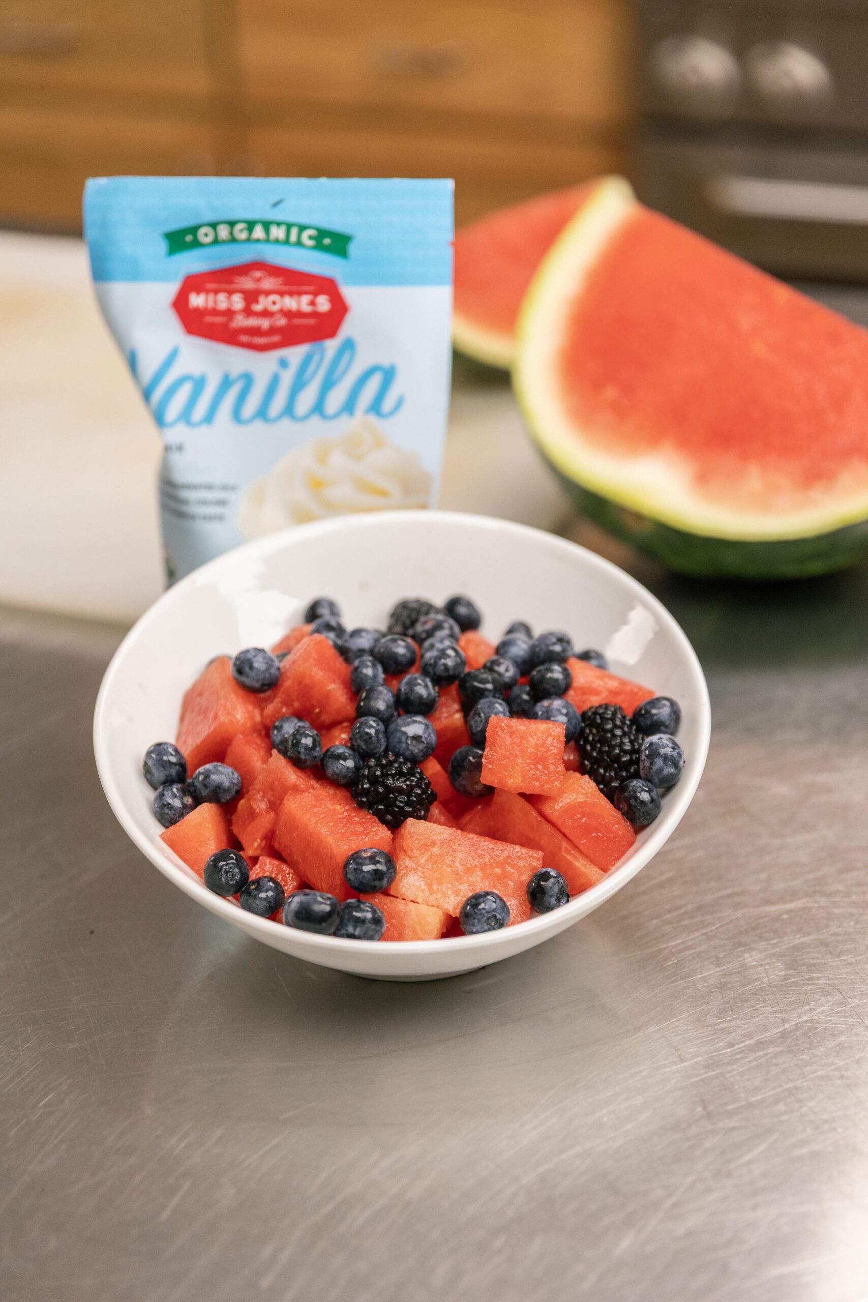 Bowl of fresh watermelon cubes, blueberries, and blackberries, with a packet of Miss Jones organic vanilla frosting in the background, showcasing a colorful and healthy fruit dessert.