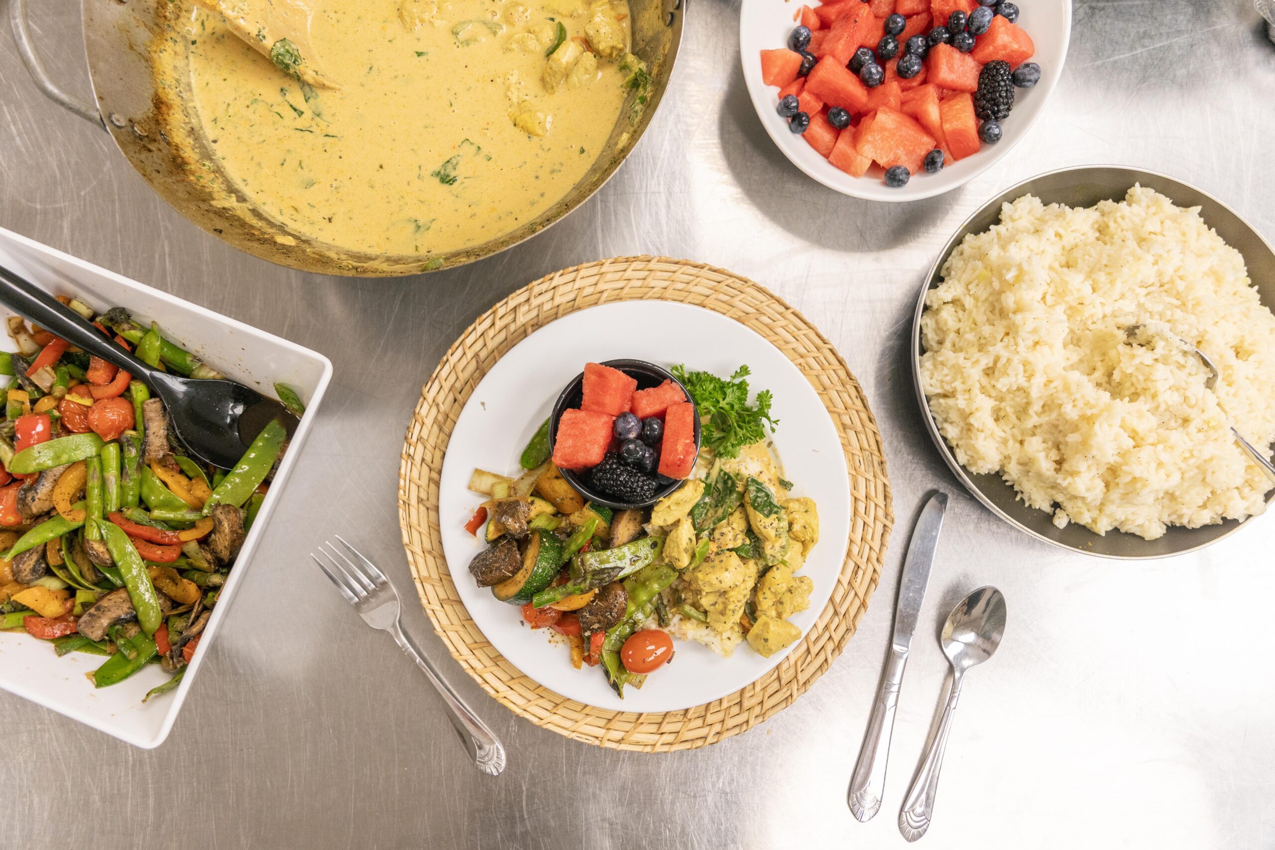 A vibrant meal featuring a plate of sautéed vegetables, chicken in curry sauce, and a side of fluffy rice, accompanied by a bowl of fresh fruit including watermelon, blueberries, and blackberries. The setting showcases a diverse and healthy dining option.