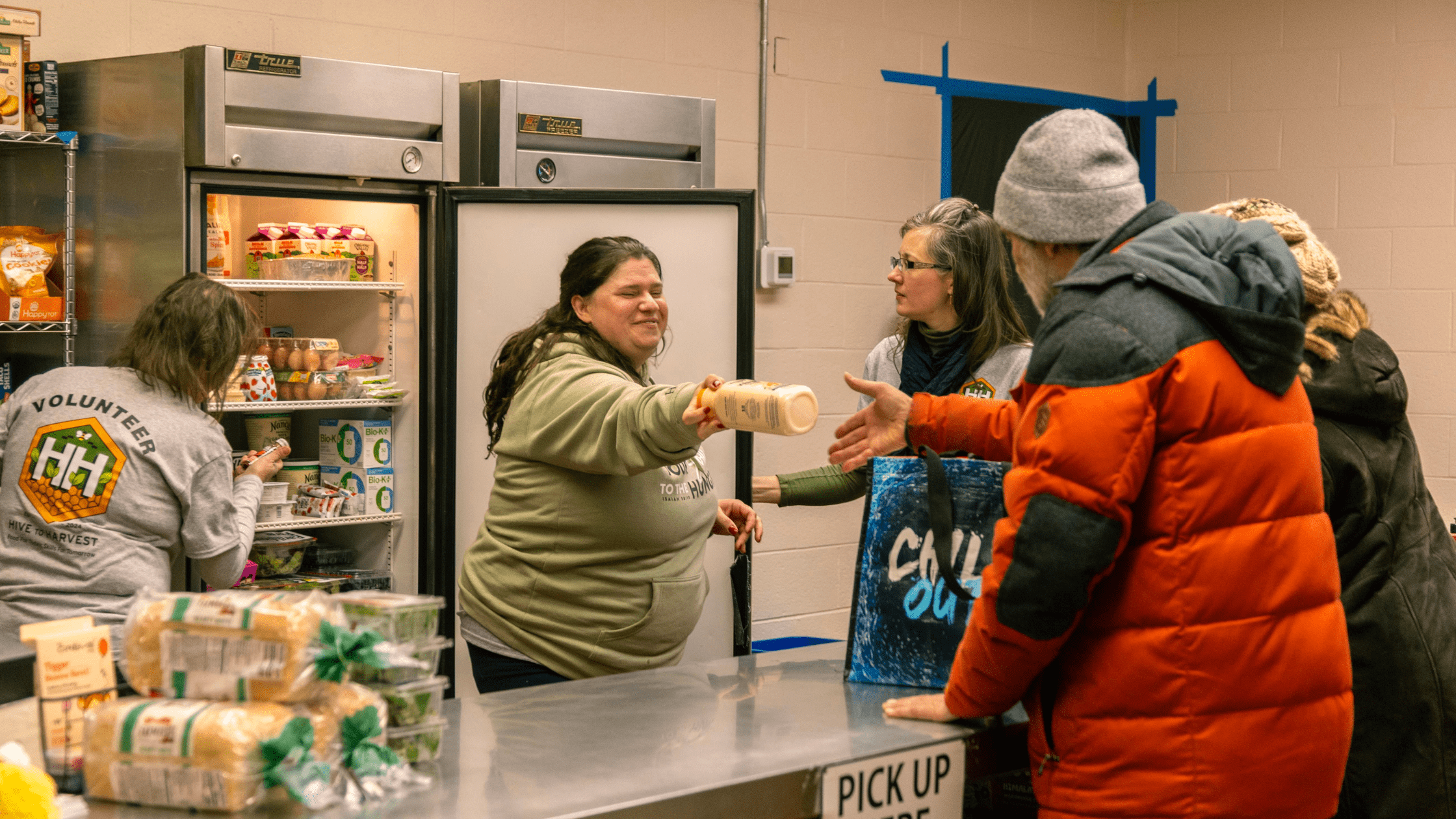 Volunteers distributing food items at a community pantry, with a woman handing a bottle to a customer while others wait in line. The scene highlights community support and food assistance efforts.