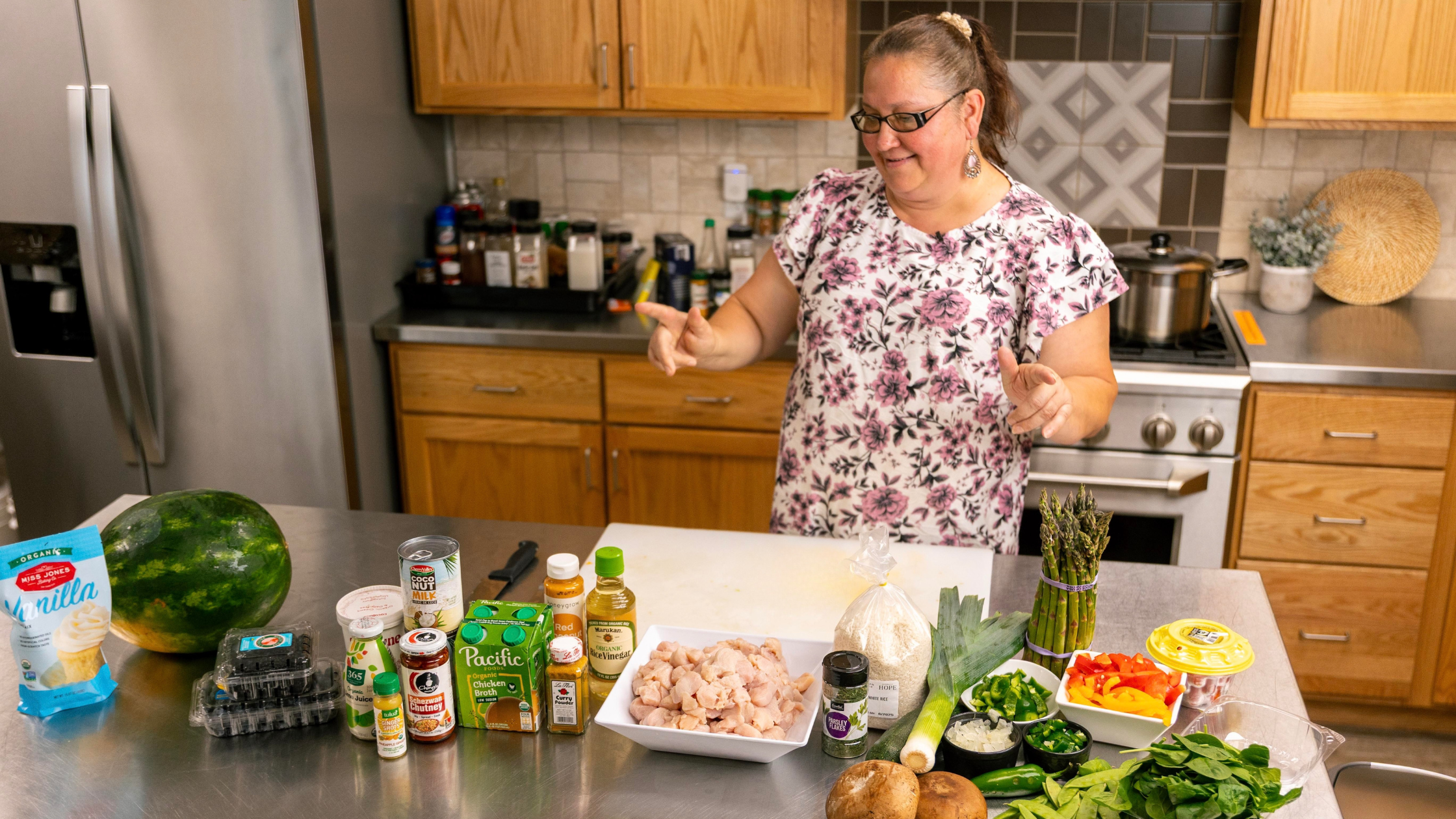 Woman in a floral shirt preparing ingredients in a kitchen, featuring chicken, vegetables, spices, and cooking supplies on a stainless steel countertop.