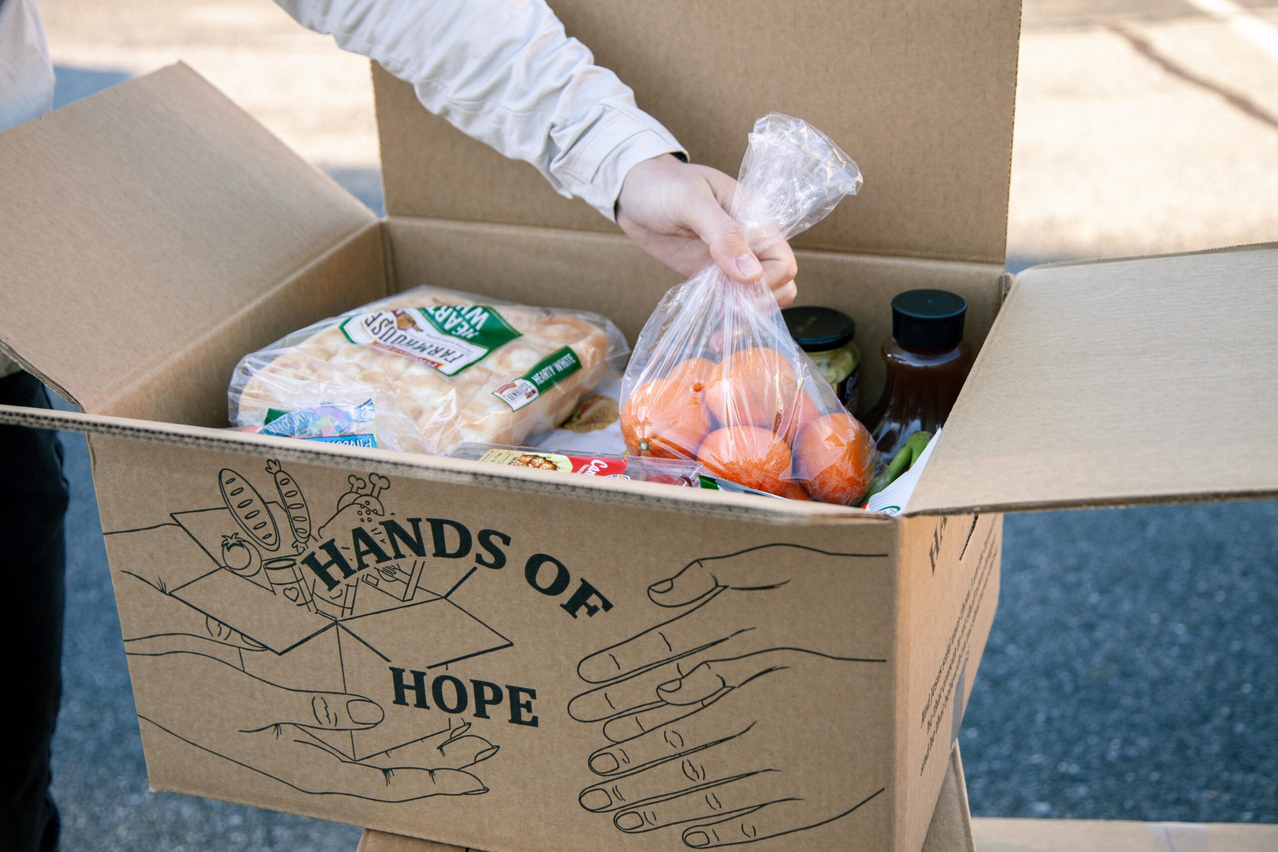 A person holding a bag of oranges while reaching into a cardboard box labeled "Hands of Hope," which contains various food items like bread, canned goods, and vegetables, highlighting community support and food distribution initiatives.