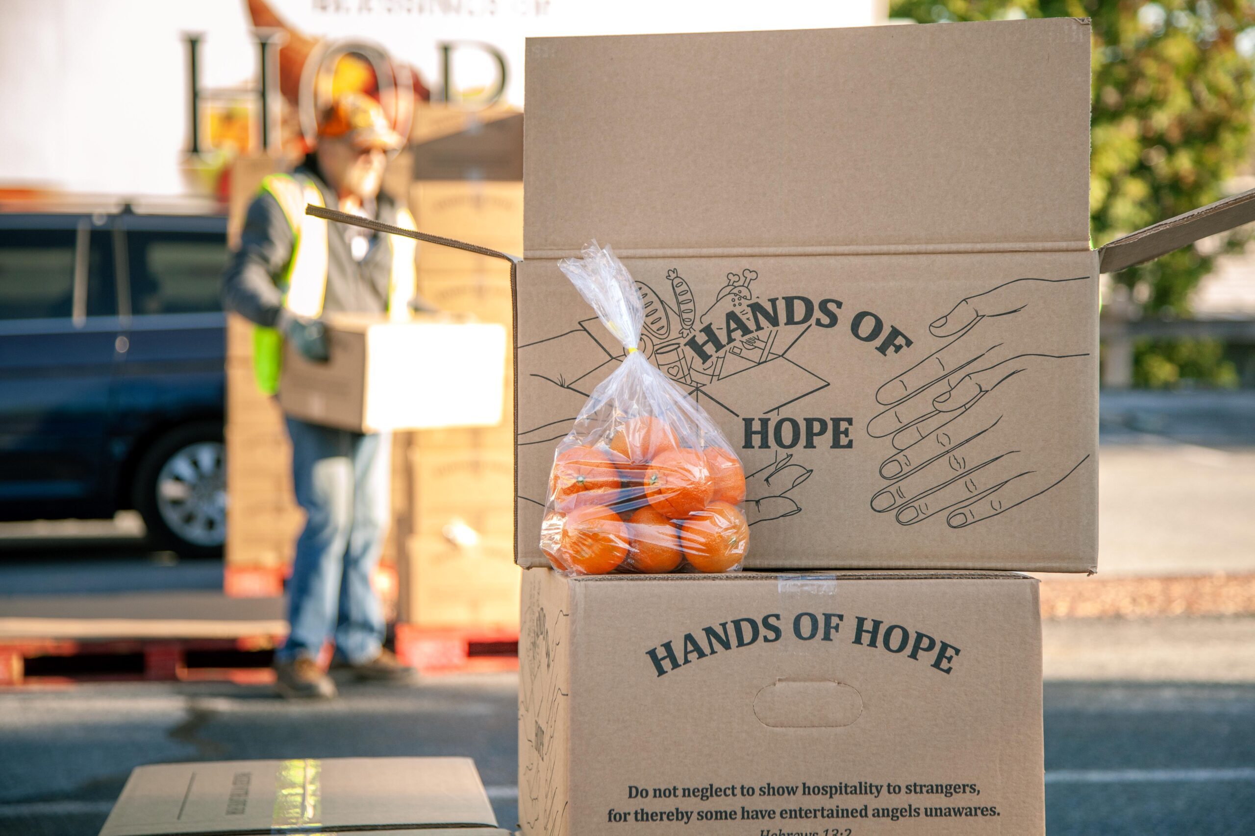 Boxes labeled "Hands of Hope" stacked at a food distribution site, with a clear plastic bag of oranges placed on top. A volunteer in a safety vest is seen in the background, carrying a box. The scene highlights community support and food assistance efforts.