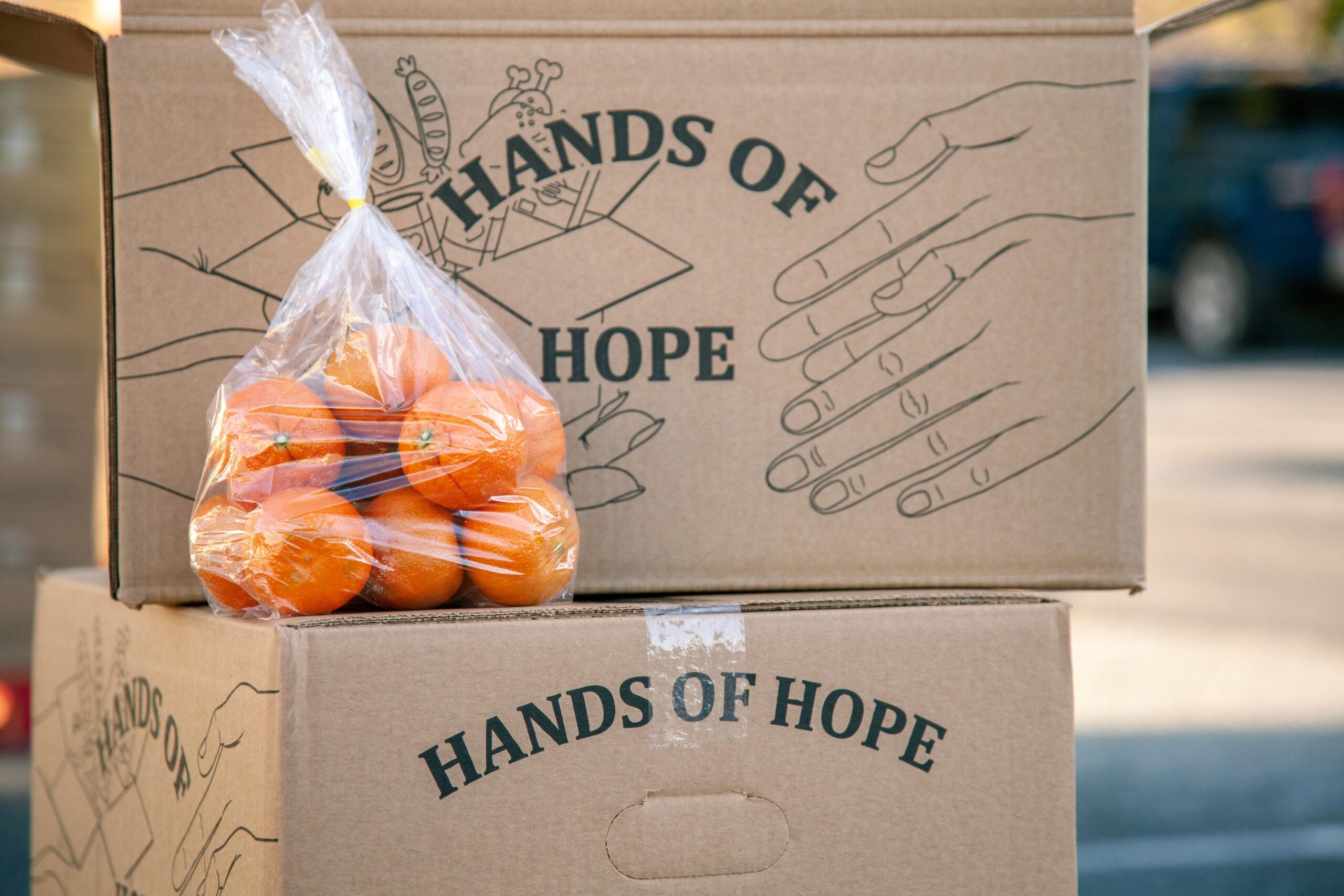 Boxes labeled "Hands of Hope" stacked together, with a clear bag of fresh oranges prominently displayed on top, symbolizing food assistance and community support.