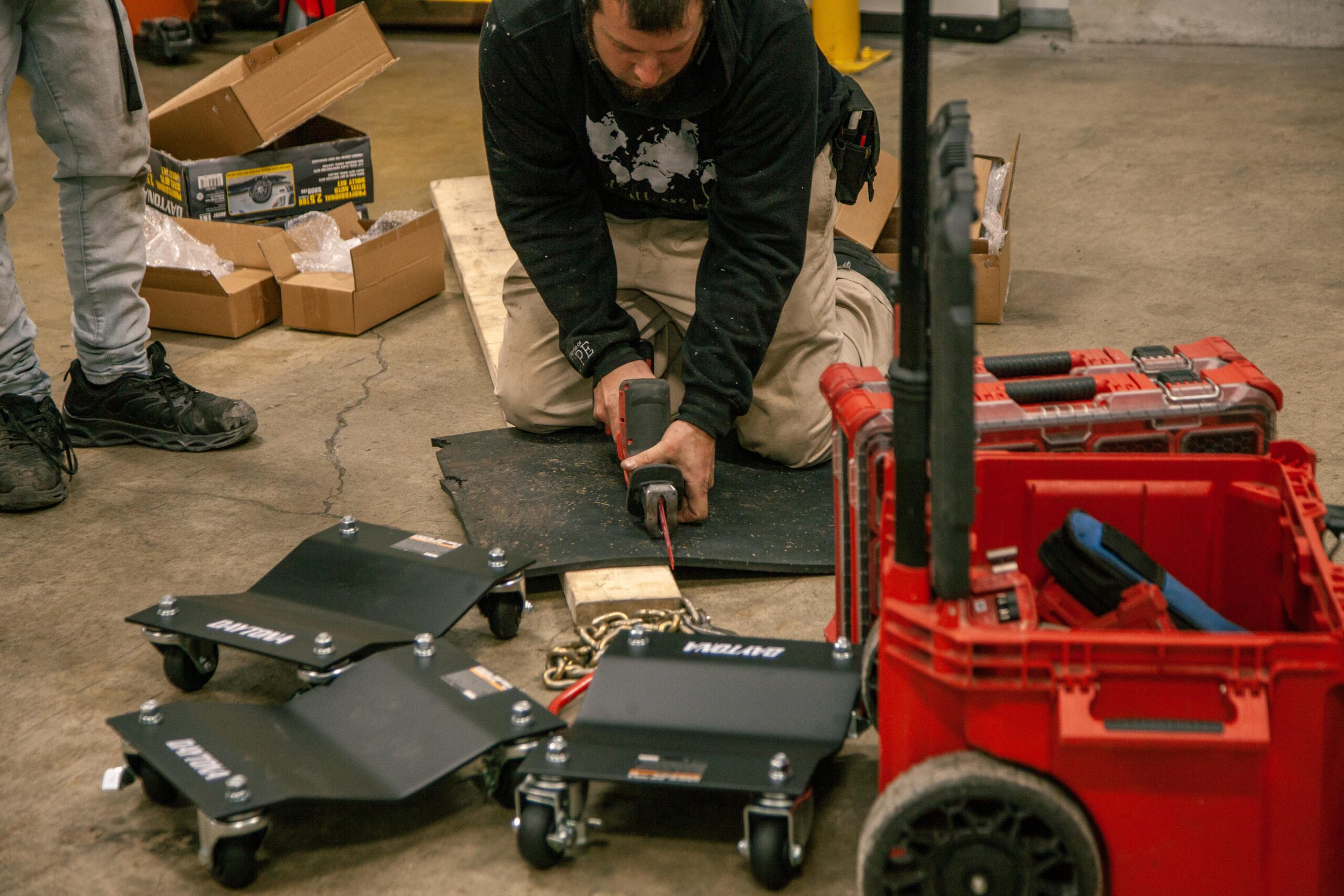 A worker using a power tool to cut a piece of material on the floor of a workshop, surrounded by tools and equipment including a red tool cart and several boxes.