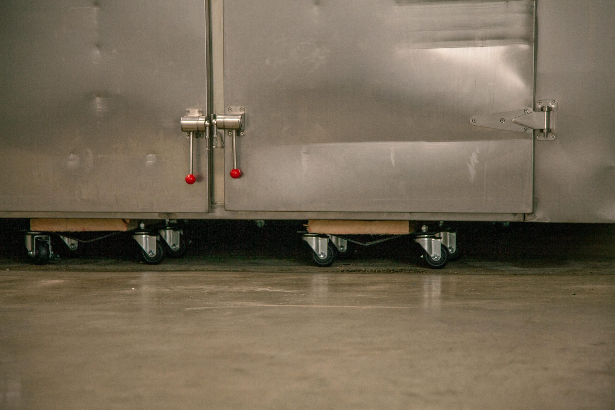 Stainless steel commercial refrigerator with red handles and wheels on a concrete floor, showcasing mobility and industrial design.