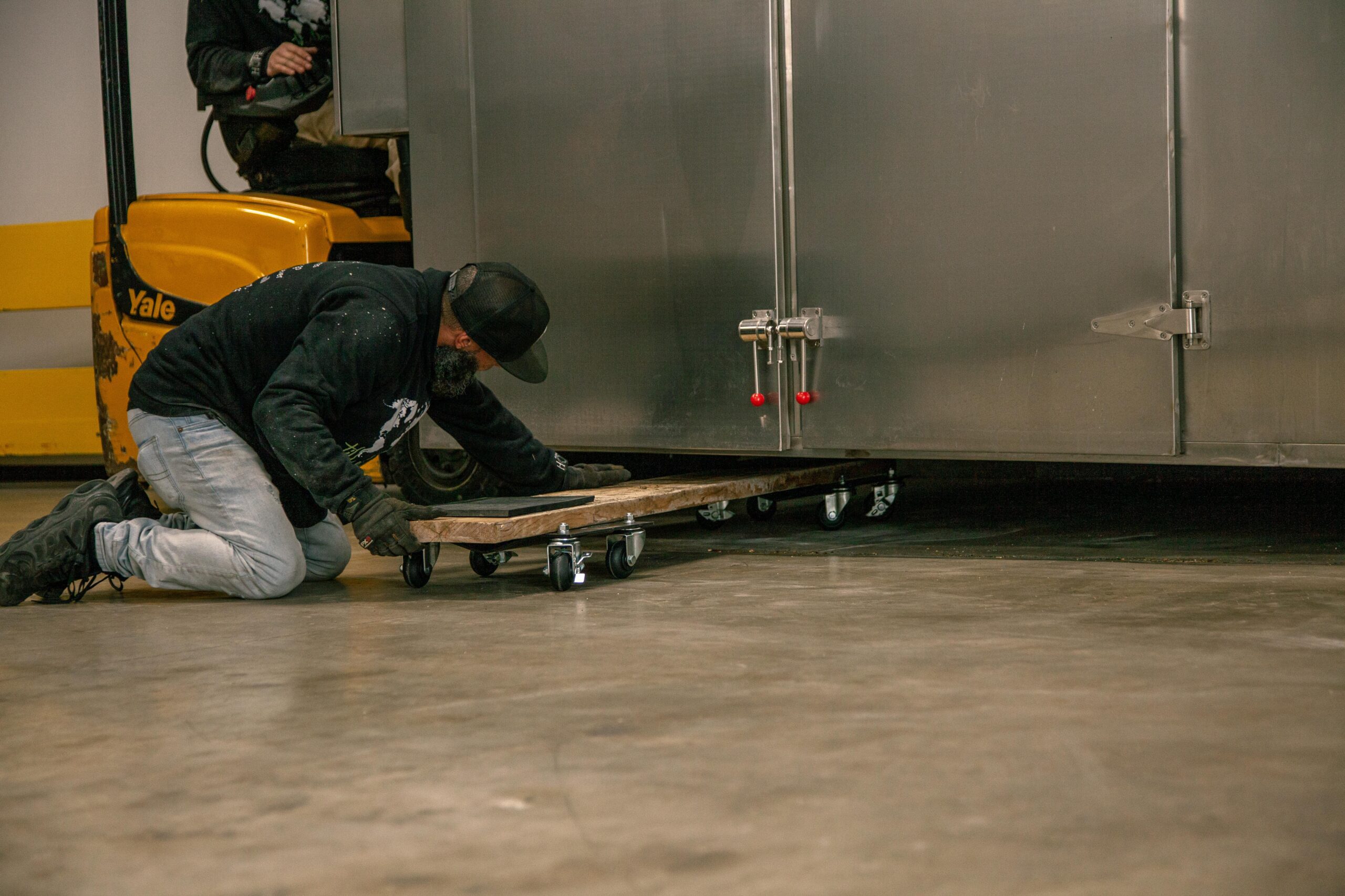 A worker in a black hoodie kneels on the warehouse floor, adjusting a wooden dolly with wheels under a stainless steel storage unit, while a yellow forklift is visible in the background.