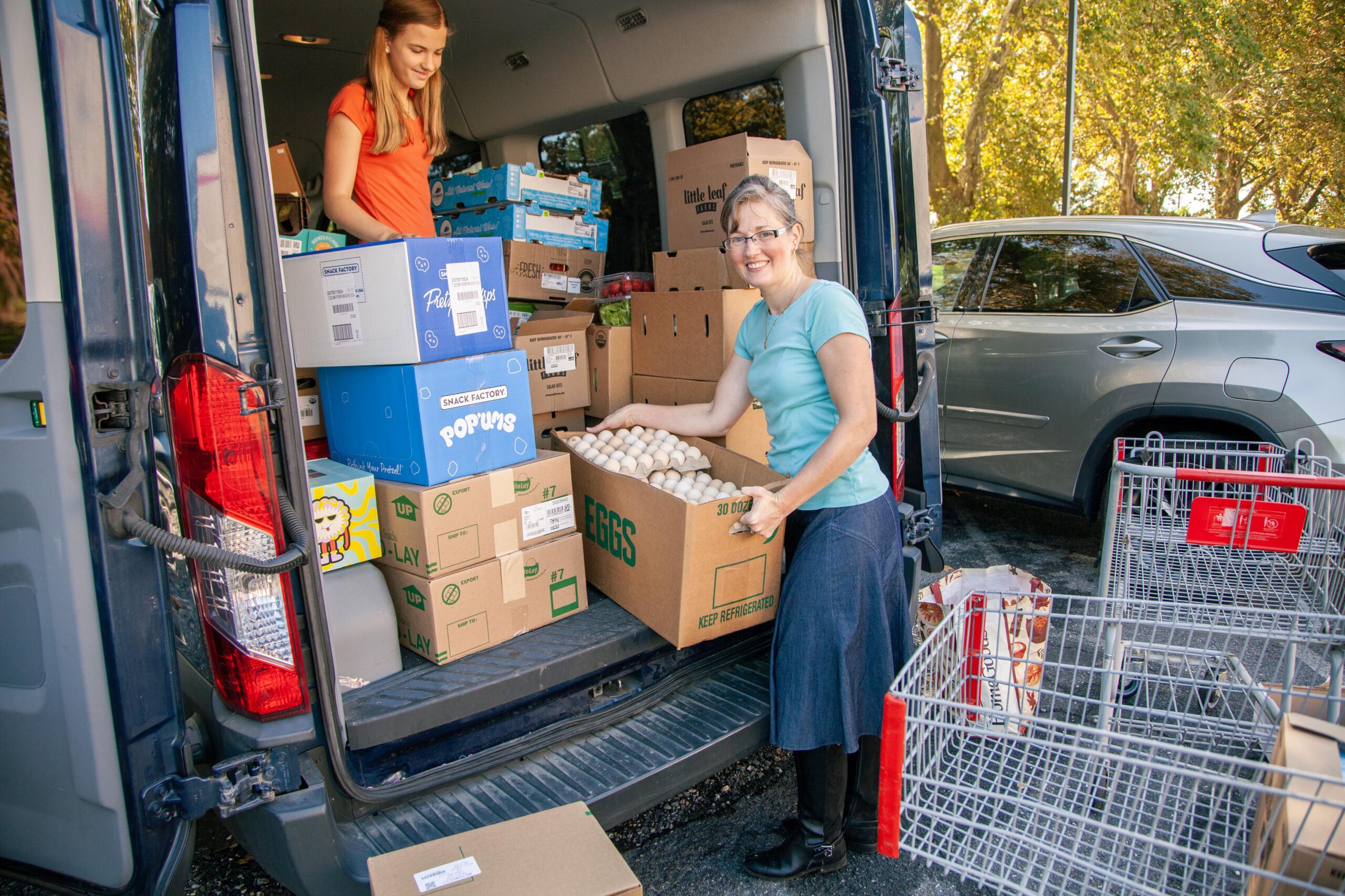 Woman unloading food boxes, including eggs and snacks, from a delivery van in a community service setting, with another person organizing items in the background.