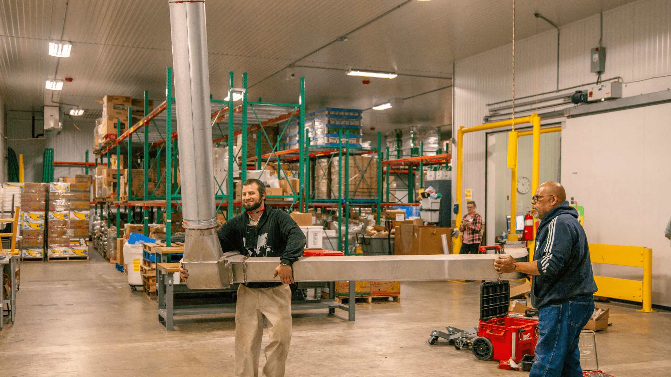 Two workers are lifting a large metal duct in a warehouse filled with shelves of boxes and supplies. The setting includes industrial shelving and various tools, illustrating a busy operational environment.