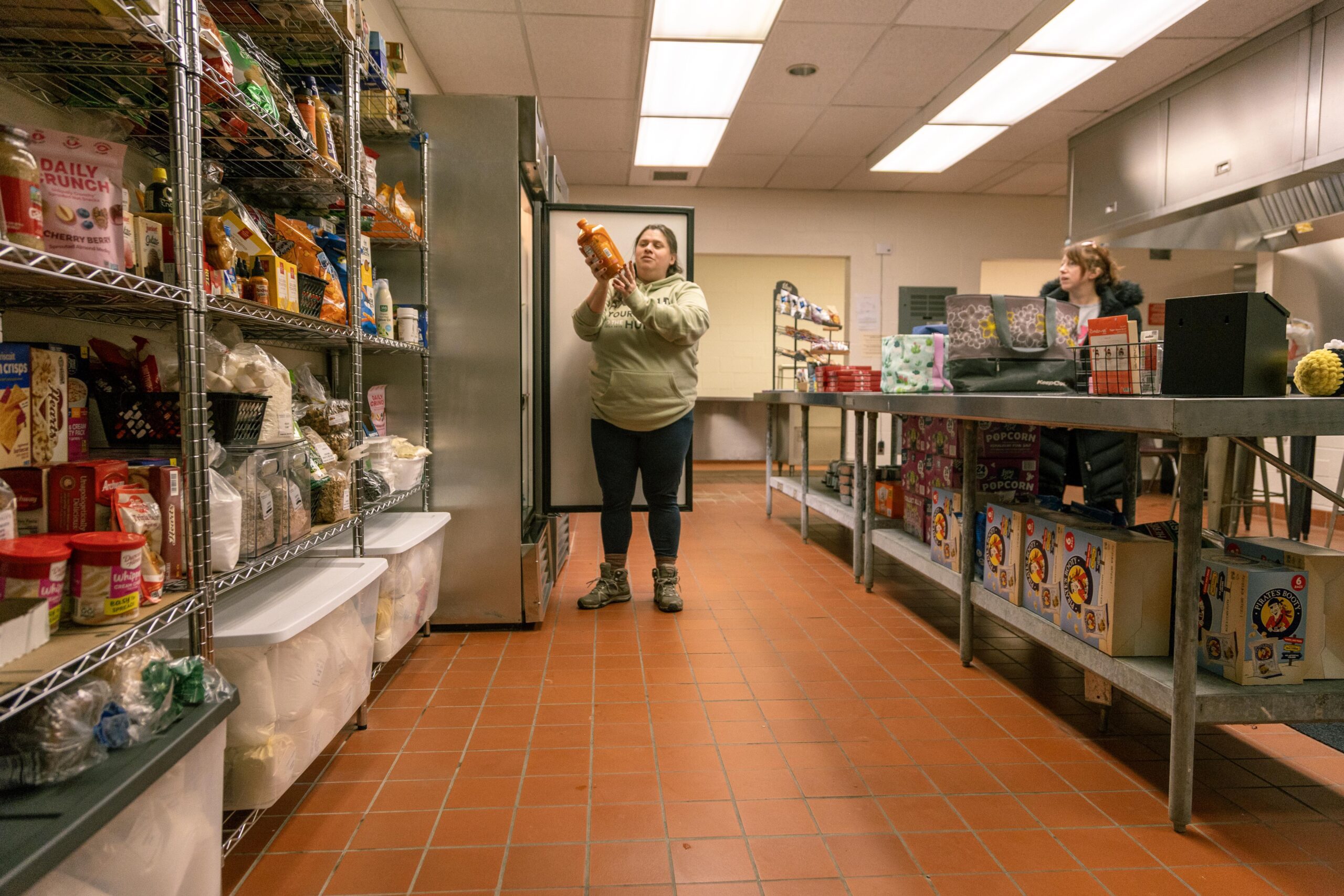 Woman organizing supplies in a well-stocked kitchen pantry with shelves filled with various food items and a refrigerator in the background. Another person is seen at a table with additional food products.