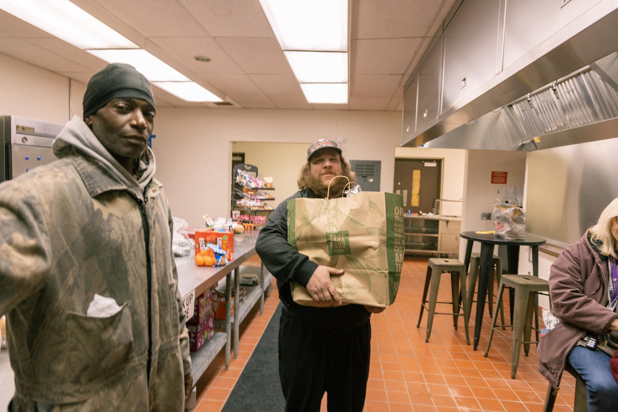 Two men and a woman inside a community kitchen, with one man holding a grocery bag. The kitchen features a countertop with snacks and fruits, and stools are visible in the background. The scene highlights food distribution efforts in a supportive environment.