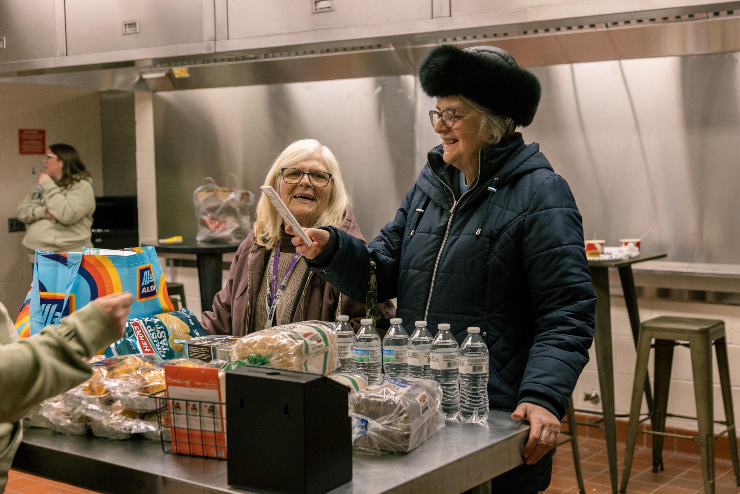 Volunteers engaging in a food distribution event, smiling and interacting at a table filled with groceries, including bread and water bottles, in a community kitchen setting.