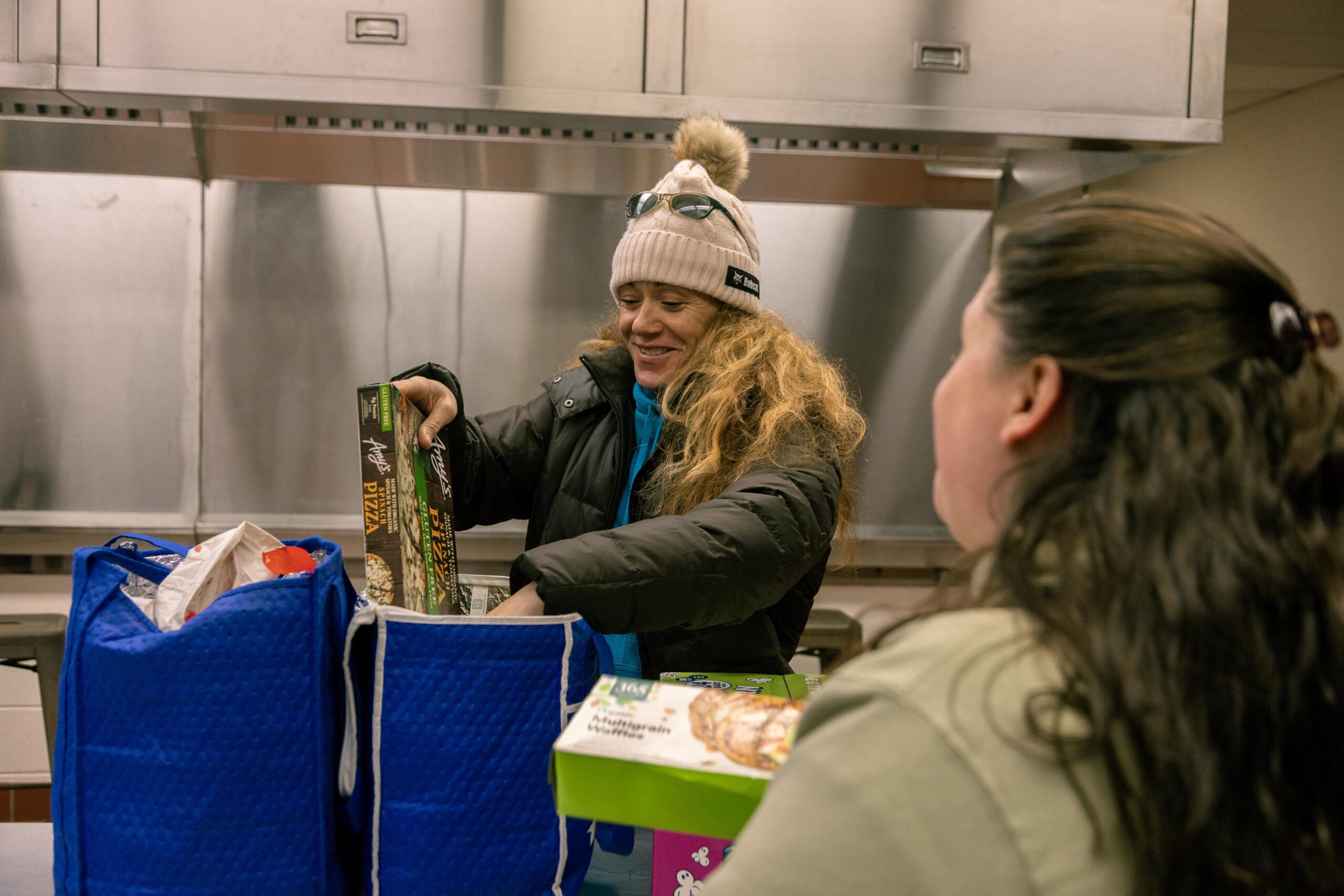 Two women are interacting in a kitchen setting, with one woman smiling while sorting through food items in blue bags. The other woman is observing her, showcasing a moment of community engagement or food distribution.