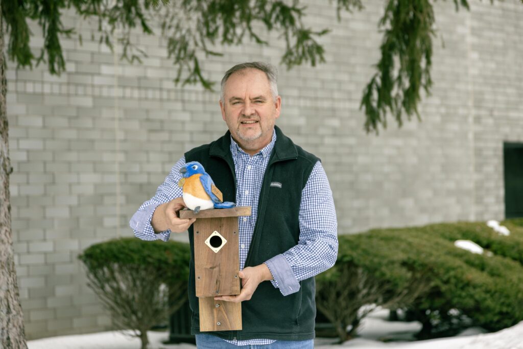 Man holding a wooden birdhouse with a plush bluebird perched on top, standing outdoors in front of a brick wall and greenery, showcasing bird conservation efforts.