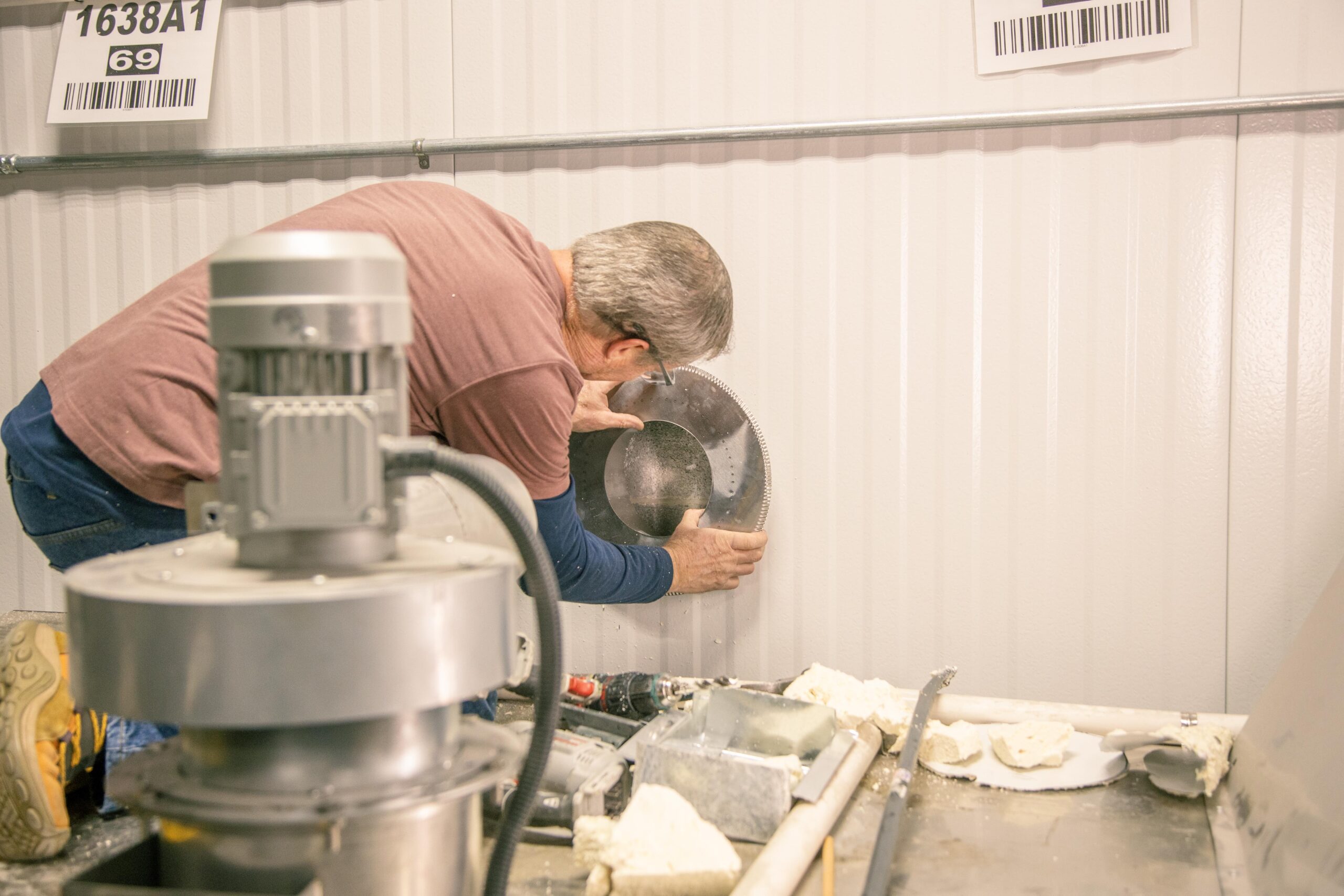 A technician installing a circular metal component on a wall in a workshop, with tools and materials scattered on the floor, showcasing a hands-on approach to equipment maintenance.