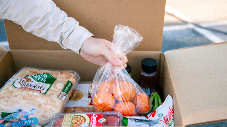 A person holding a bag of oranges while unpacking a cardboard box filled with groceries, including bread, sauces, and vegetables.