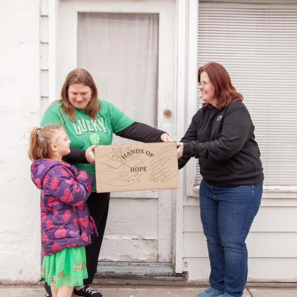 A woman in a green shirt and a woman in a black hoodie are handing a box labeled "HANDS OF HOPE" to a young girl in a purple jacket and green dress outside a house.
