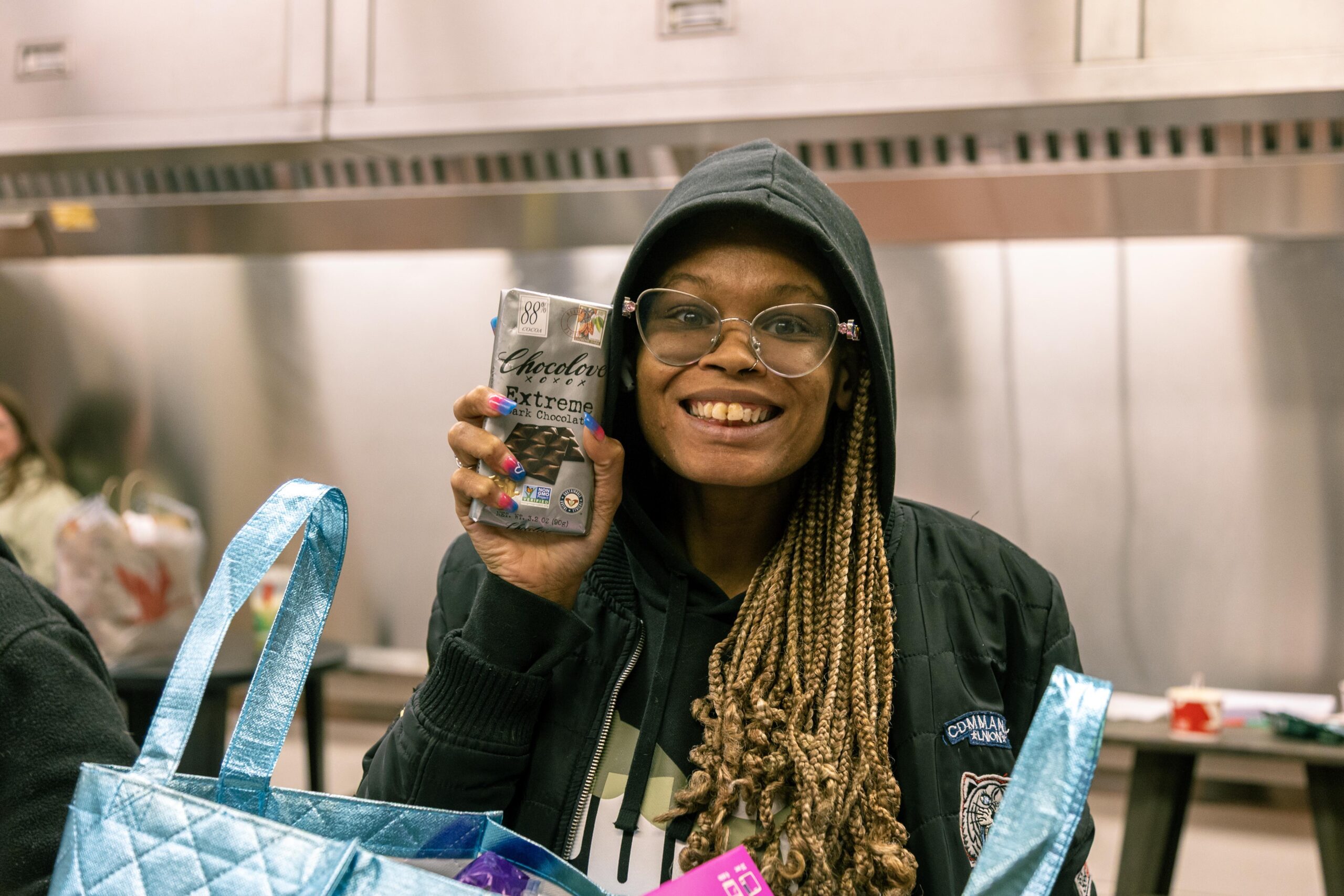 Smiling woman holding a bar of Chocolove Extreme dark chocolate in a kitchen setting, wearing glasses and a black hoodie, with a blue tote bag nearby filled with various items.