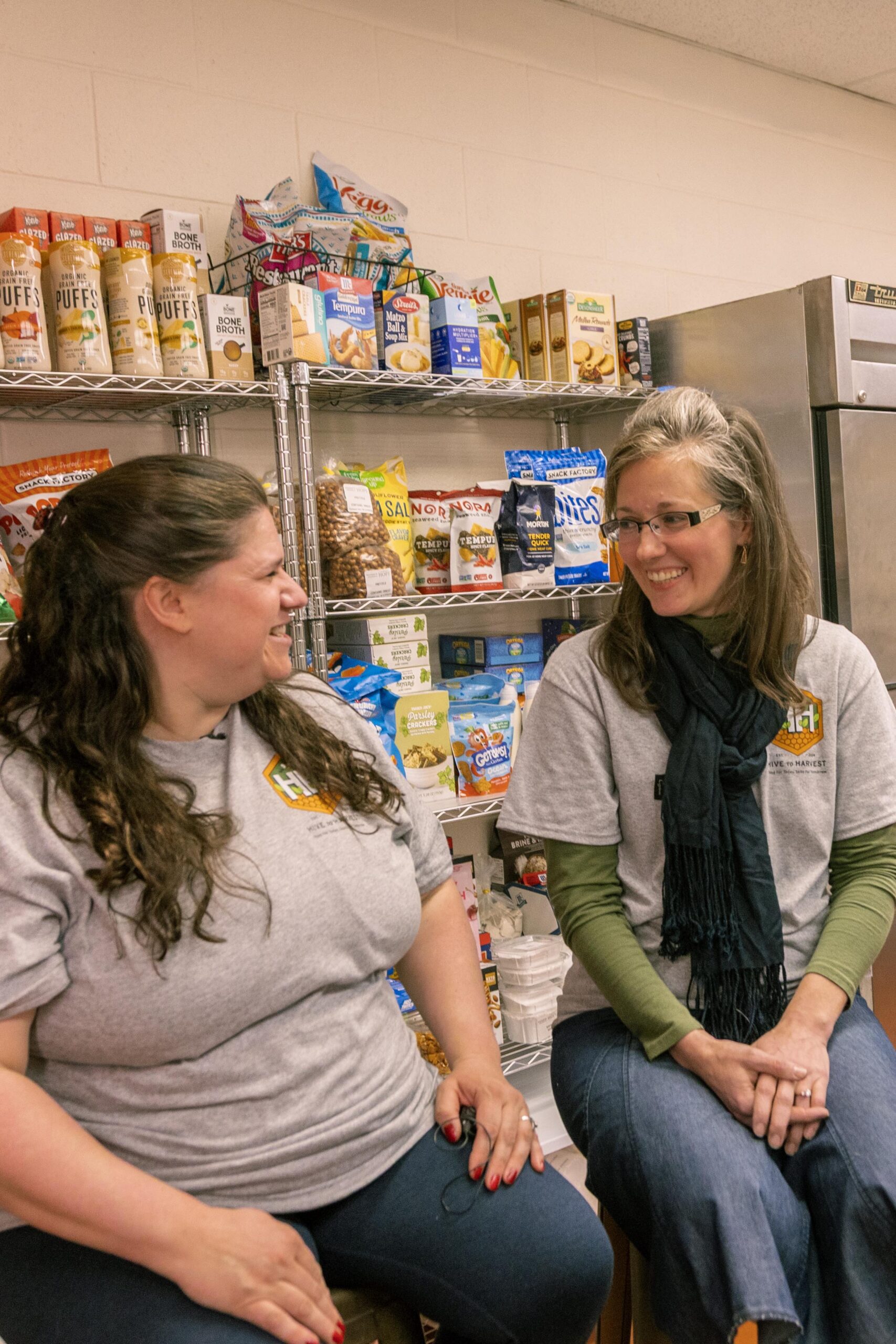 Two women wearing gray t-shirts sit together and smile in a food pantry setting, with shelves stocked with various snacks and food items in the background.