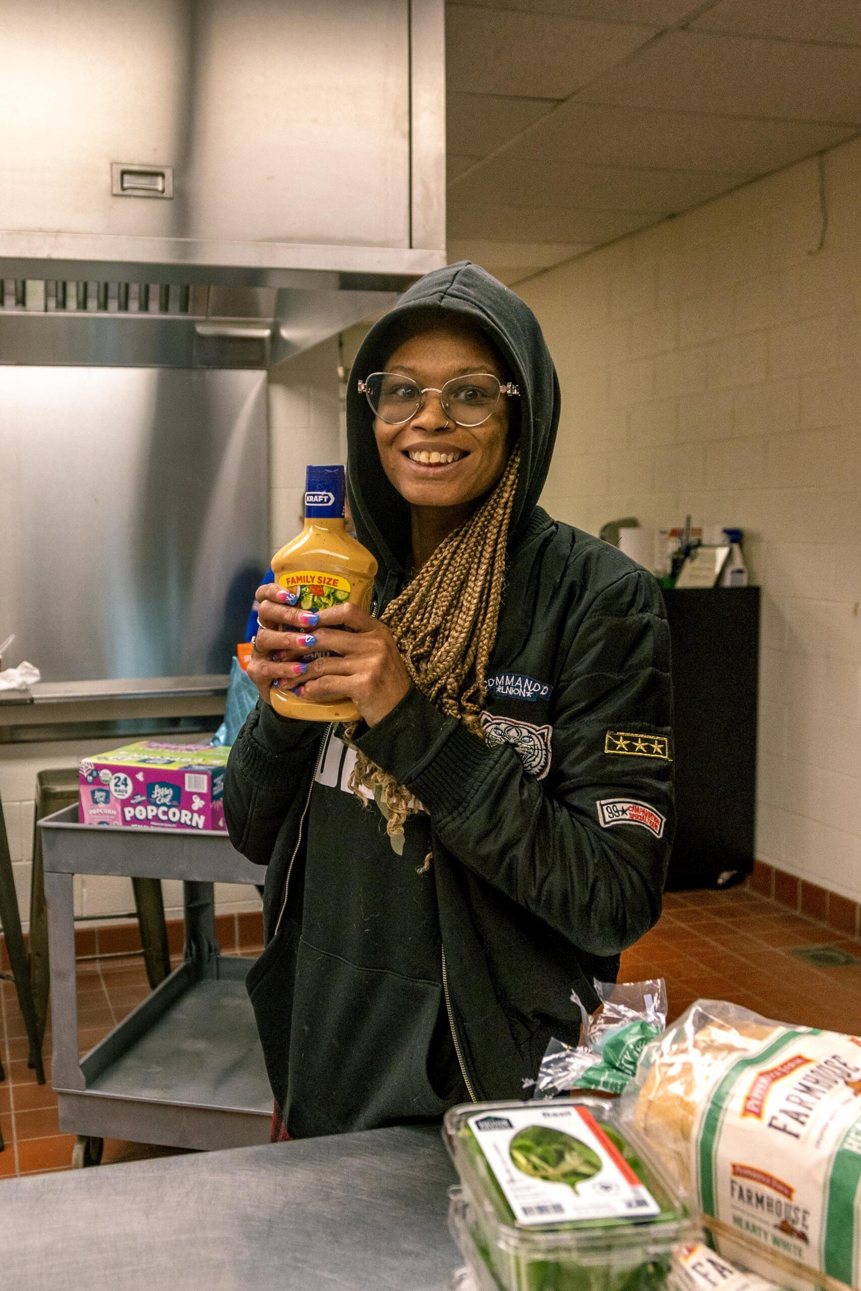 Smiling individual holding a bottle of mustard in a kitchen setting, with various food items on a table, including bread and vegetables, showcasing a warm and inviting atmosphere.