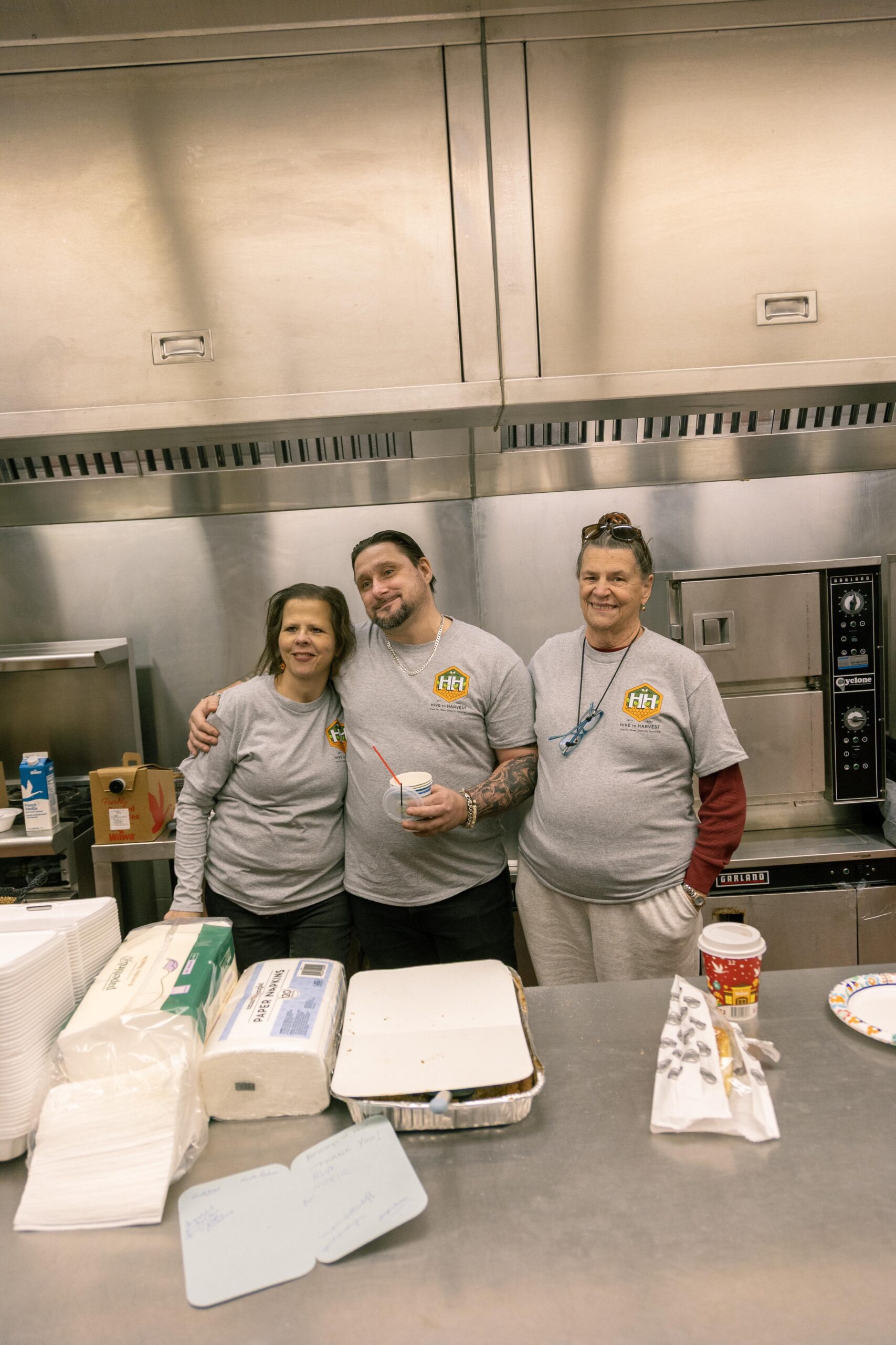 Three volunteers in gray t-shirts stand together in a kitchen, smiling and posing for the camera. They are surrounded by food supplies, including disposable containers and drinks, highlighting their involvement in a community service or food preparation event.