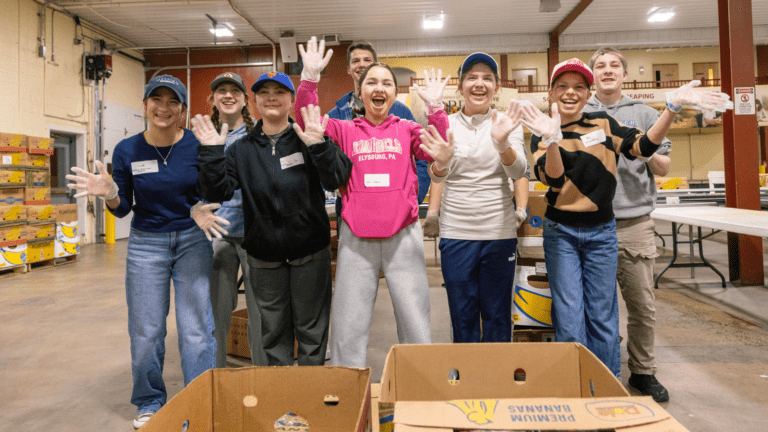 Group of volunteers smiling and waving at a food distribution center, wearing gloves and casual clothing, with cardboard boxes in the foreground. The setting highlights community service and teamwork in a supportive environment.