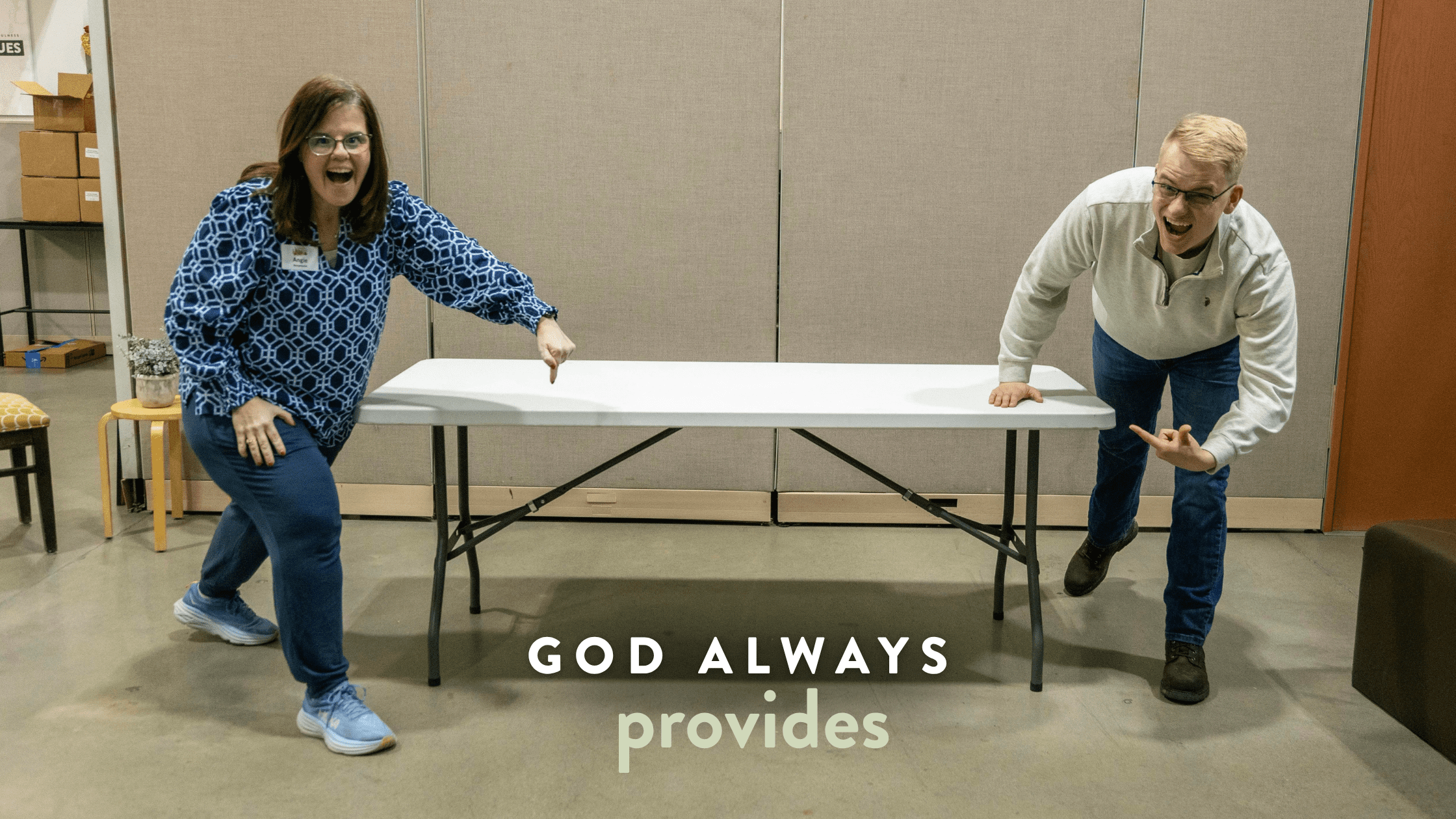 Two enthusiastic individuals point at a white folding table in a community space, conveying a message of positivity with the text "God Always Provides" displayed prominently. The setting features a neutral background with boxes and chairs, emphasizing a welcoming environment.