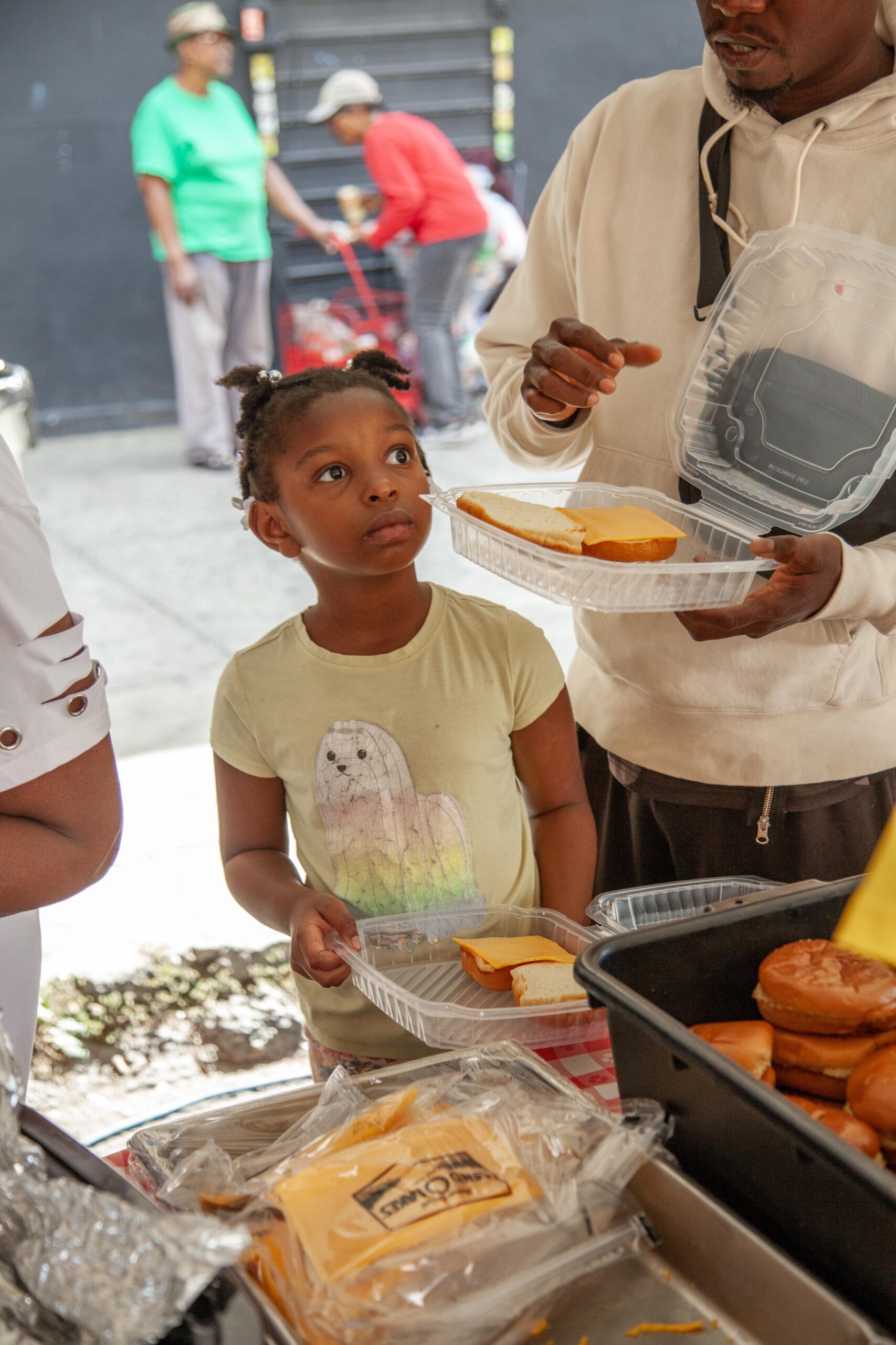 Young girl with braided hair looks up curiously while holding a plastic tray with a sandwich, as an adult hands her a sandwich. In the background, other people are gathered, some enjoying food at a community event.