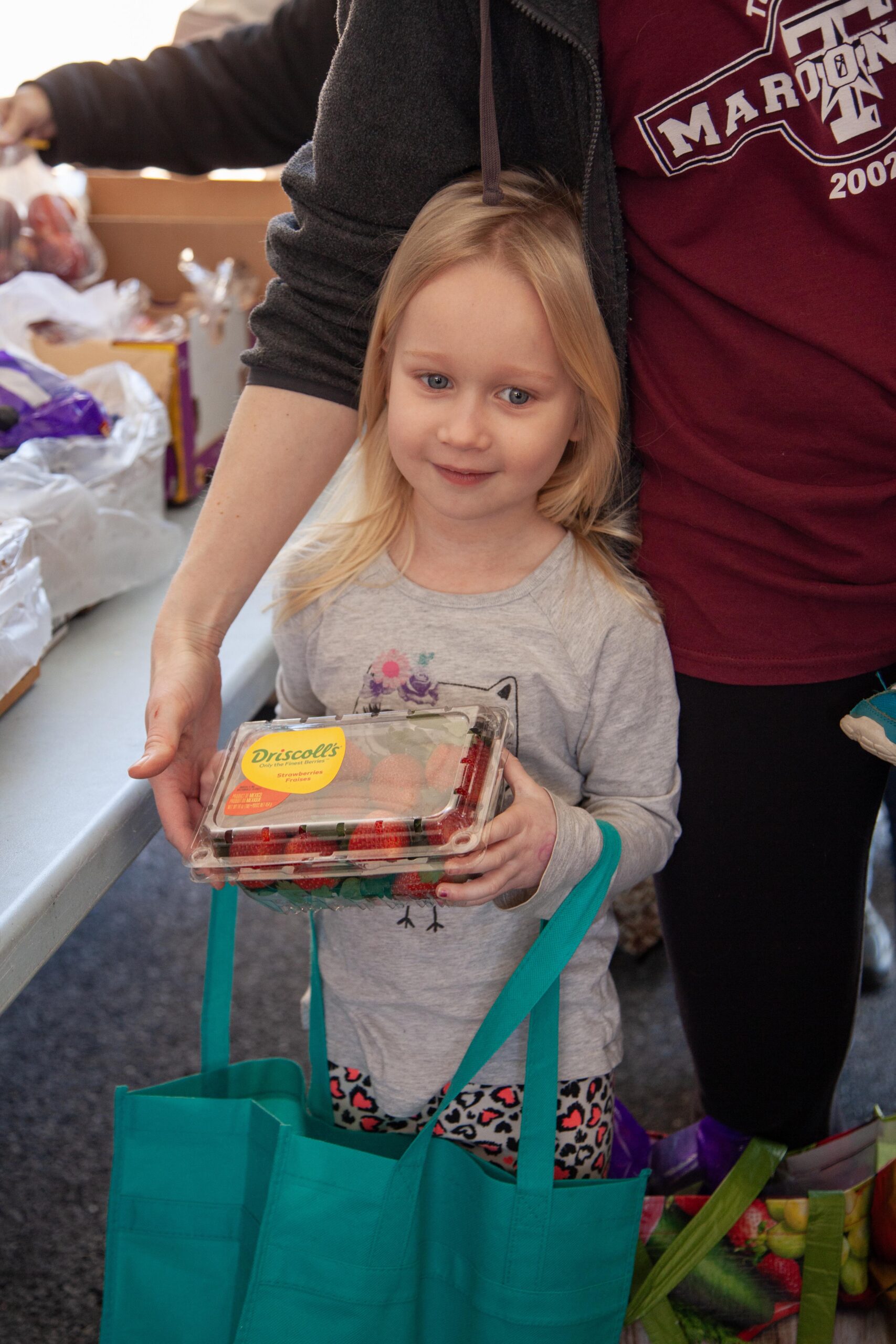 Young girl holding a container of fresh strawberries while standing next to an adult, with colorful reusable bags in a community food distribution setting.