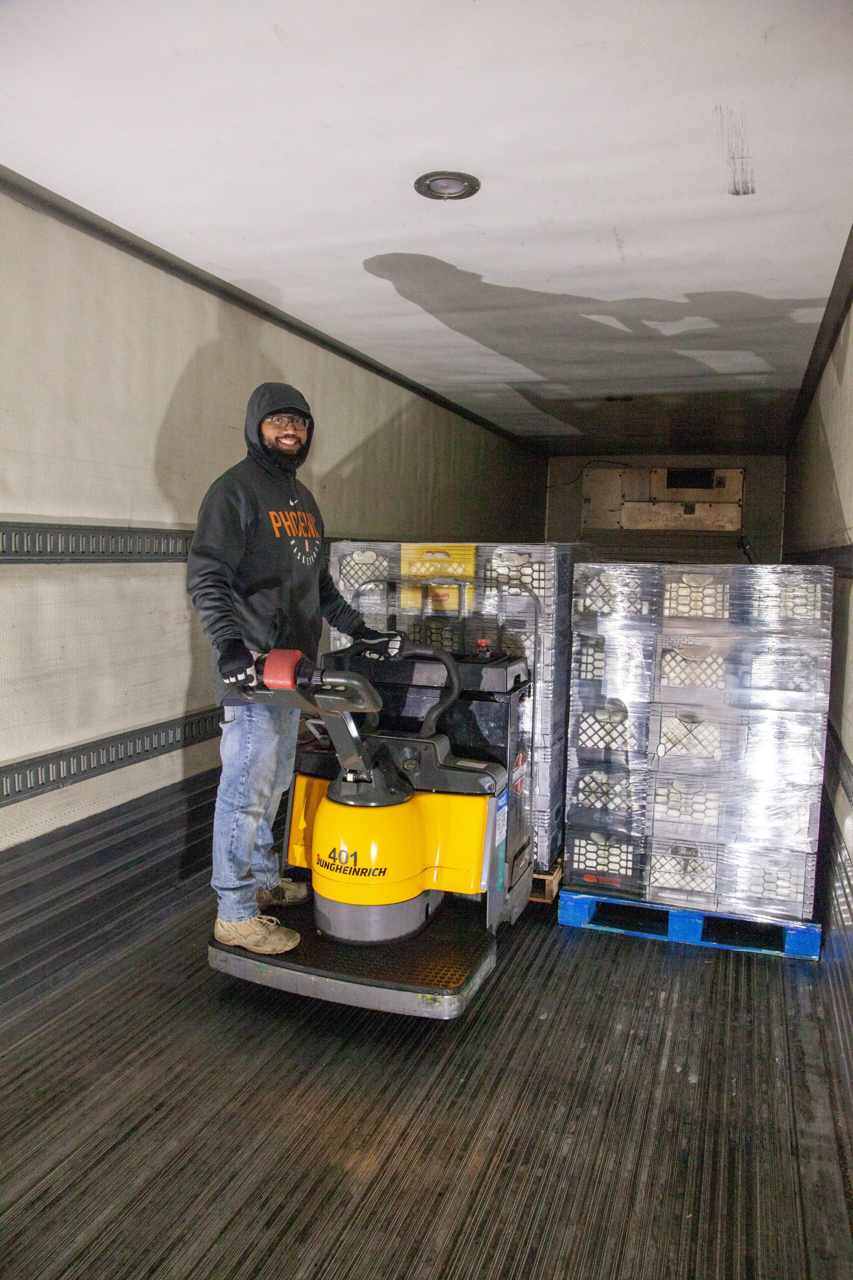 Warehouse worker operating a powered pallet jack inside a delivery truck, surrounded by stacked pallets and crates. The setting highlights logistics and material handling in a warehouse environment.