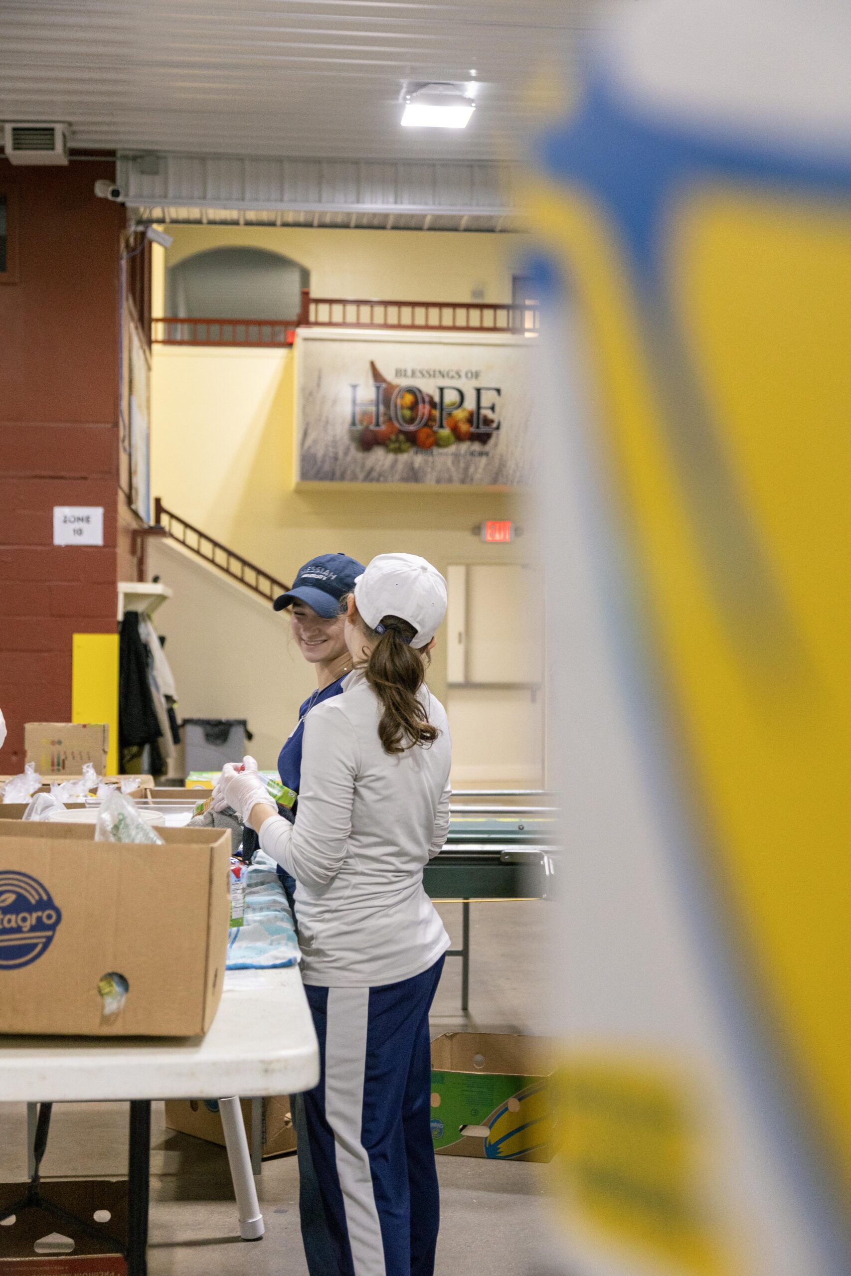 Two women volunteer at a food distribution center, preparing boxes while smiling and engaging in conversation. The background features a sign that reads "Blessings of Hope," emphasizing the community support effort.