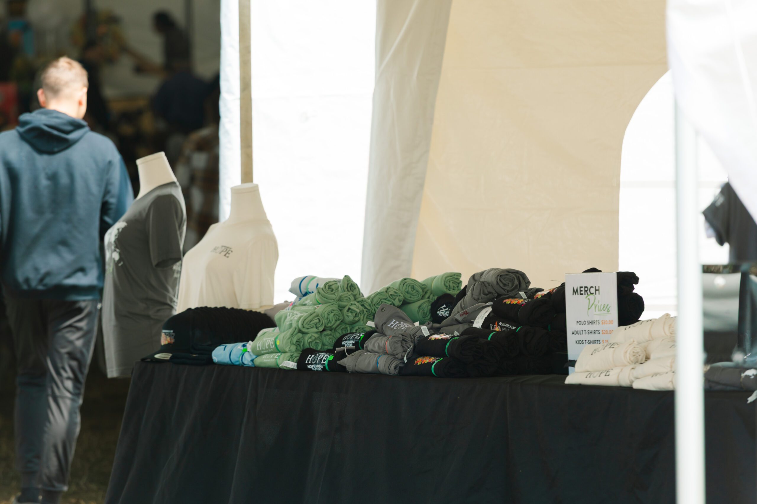 Merchandise display at an outdoor event featuring various t-shirts and polo shirts neatly arranged on a black table, with a sign listing prices for adult and kids' sizes. A person in a hoodie is seen walking in the background.