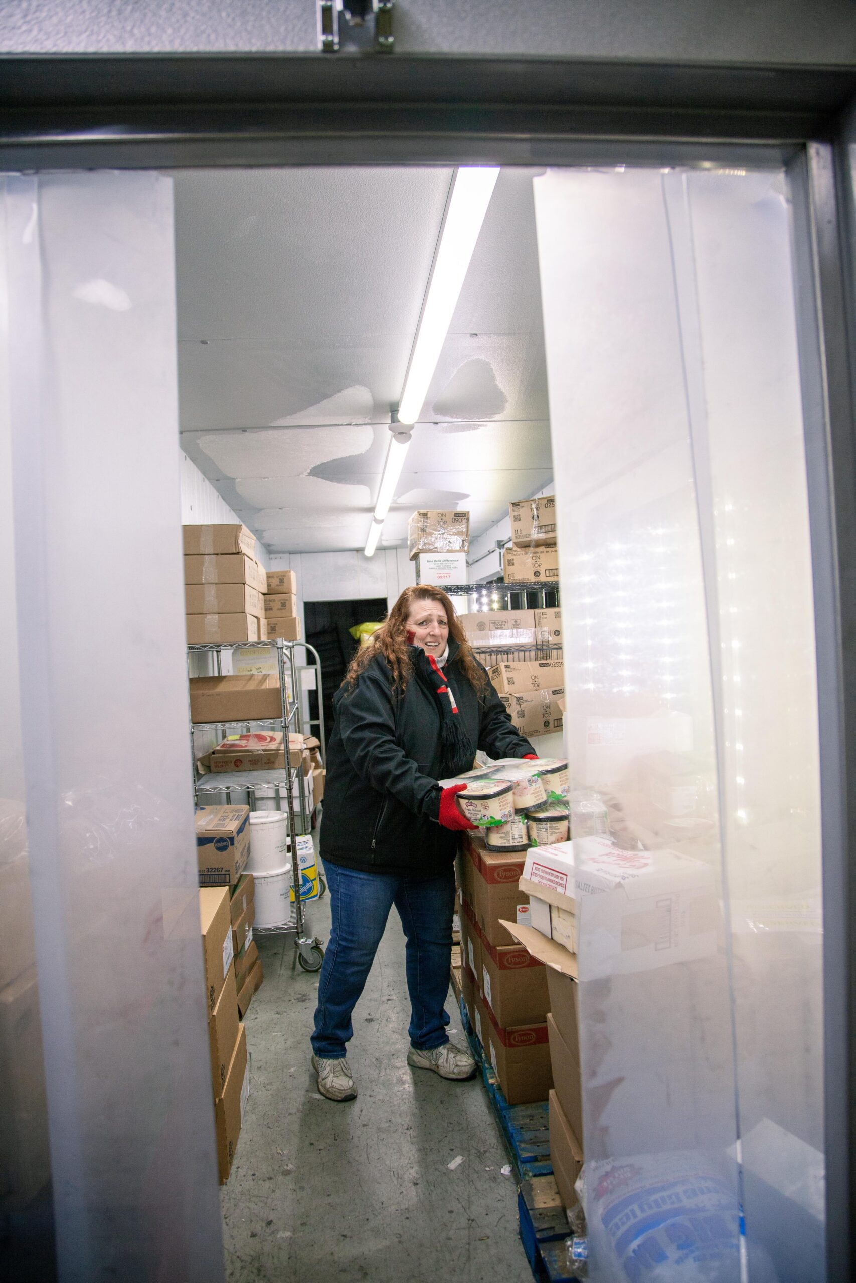 Woman wearing gloves and a black jacket carrying boxes of food inside a cold storage facility, surrounded by shelves filled with food items and cardboard boxes.