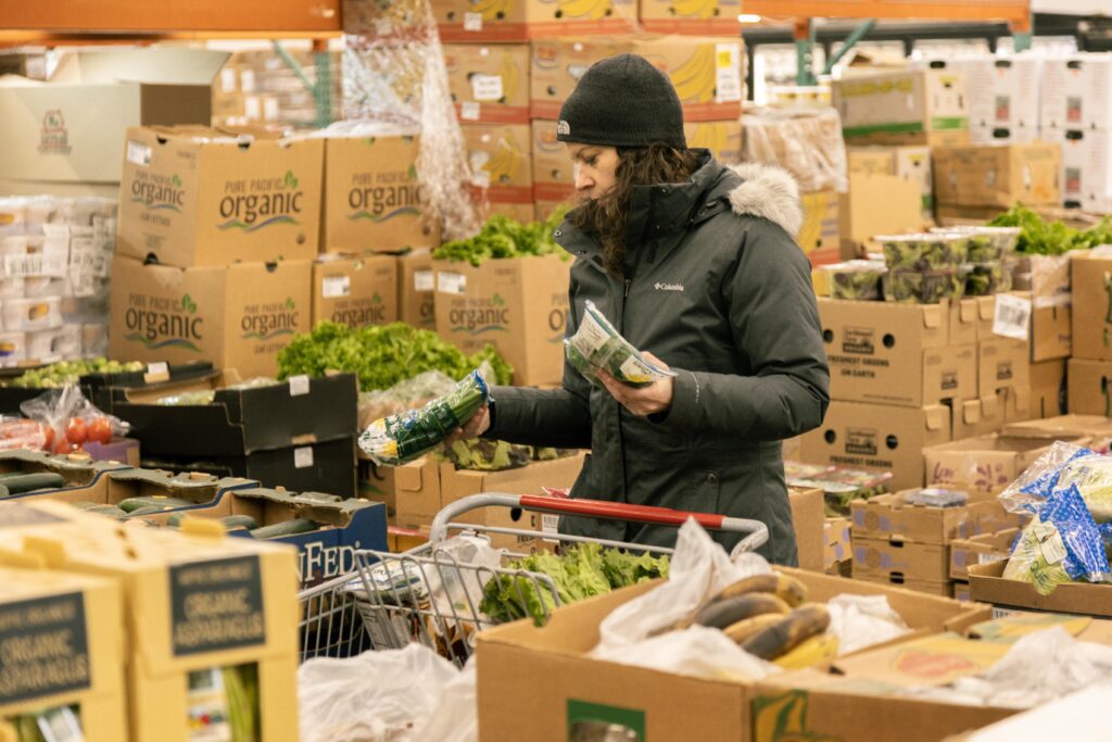 Woman shopping for organic produce in a grocery store, surrounded by boxes of fresh vegetables and fruits. She is holding two packages while examining the items. A shopping cart is nearby, filled with various greens and vegetables.