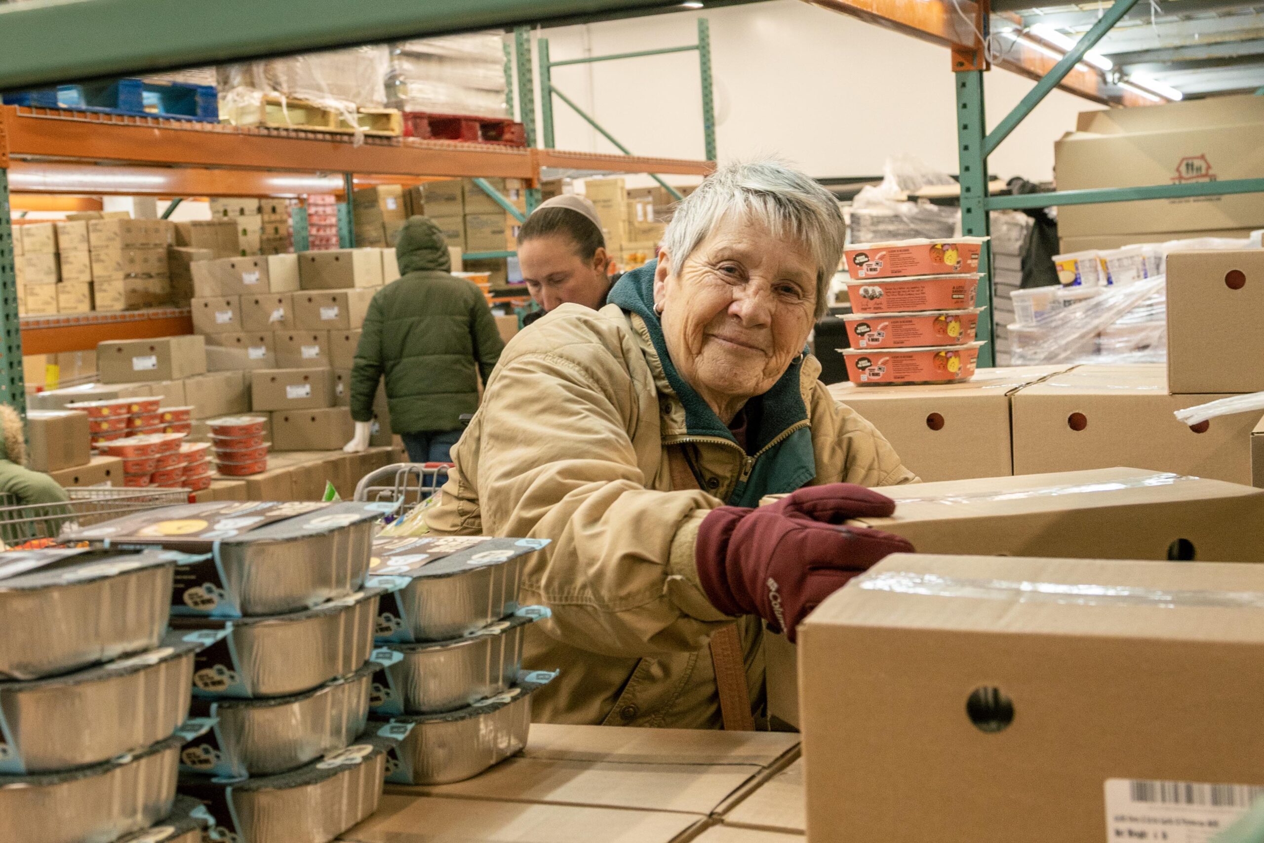 Senior volunteer organizing food boxes in a warehouse, surrounded by stacked containers and shelves filled with supplies, contributing to community support efforts.