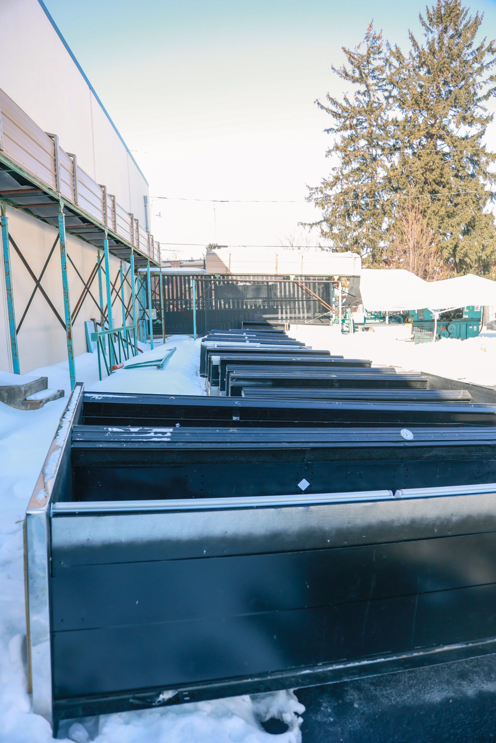 Snow-covered outdoor area featuring multiple black storage containers lined up along a concrete path, with a building and trees in the background.