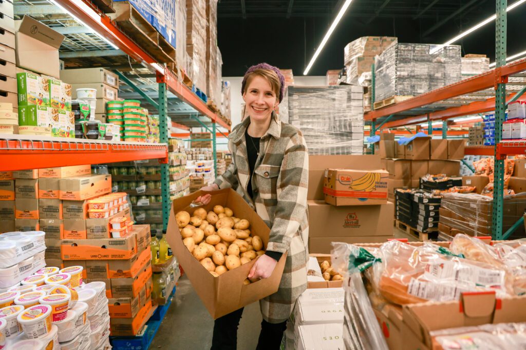 Woman holding a box of fresh potatoes in a grocery warehouse aisle, surrounded by shelves stocked with various food items and packaging.