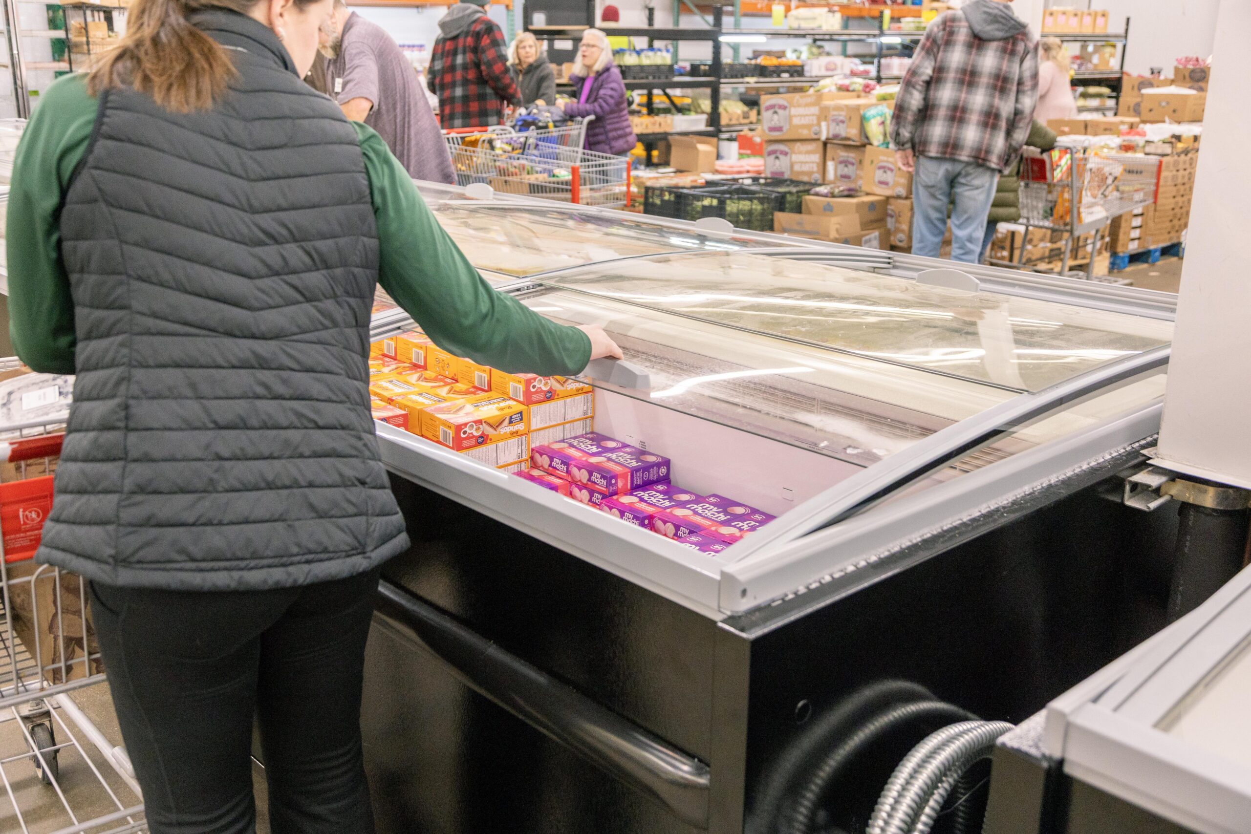 Woman selecting frozen treats from a display freezer in a grocery store, with shoppers browsing in the background.