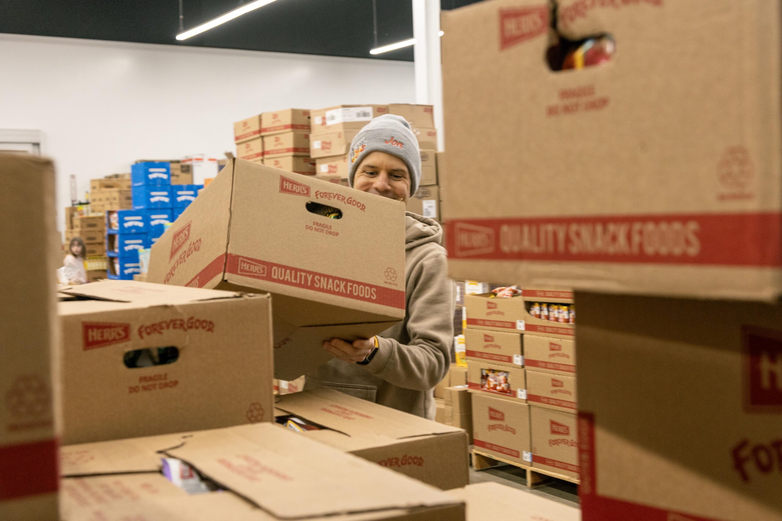 Man carrying a box of Herr's quality snack foods in a warehouse filled with stacked boxes, showcasing food distribution and storage efforts.