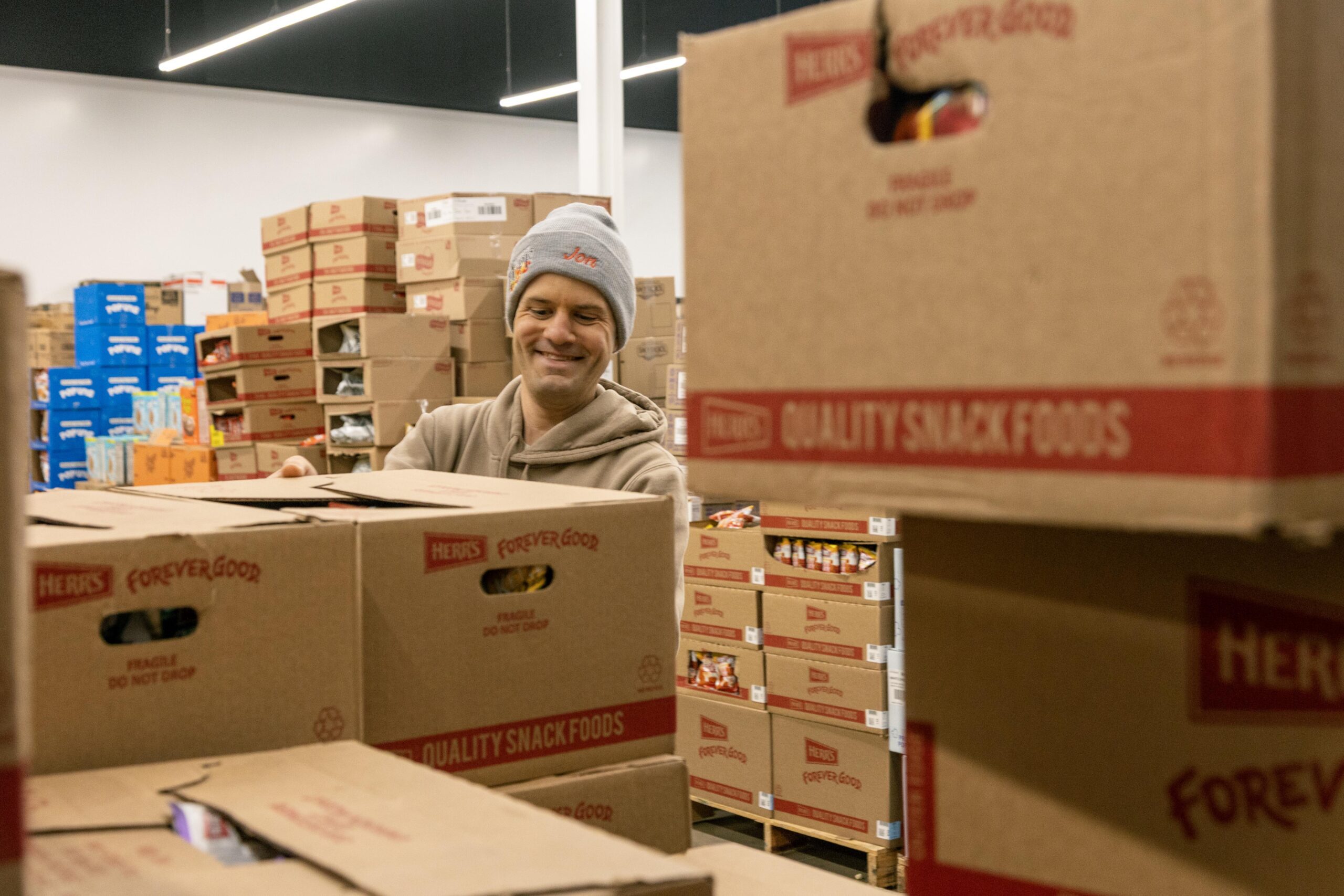 Man smiling while organizing snack food boxes in a warehouse, surrounded by stacked cardboard boxes labeled "Herr's Quality Snack Foods." The setting is bright and spacious, emphasizing the community effort in food distribution.