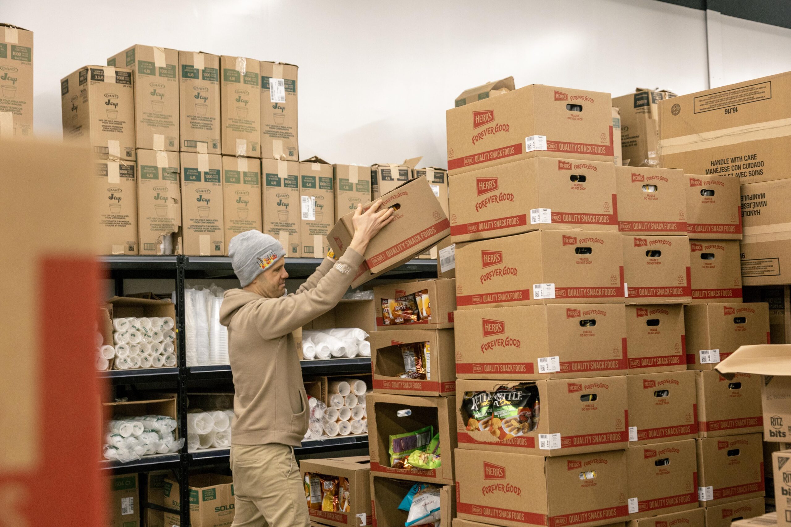 Person organizing boxes of snack foods in a storage area, surrounded by stacked cardboard boxes labeled with various products.