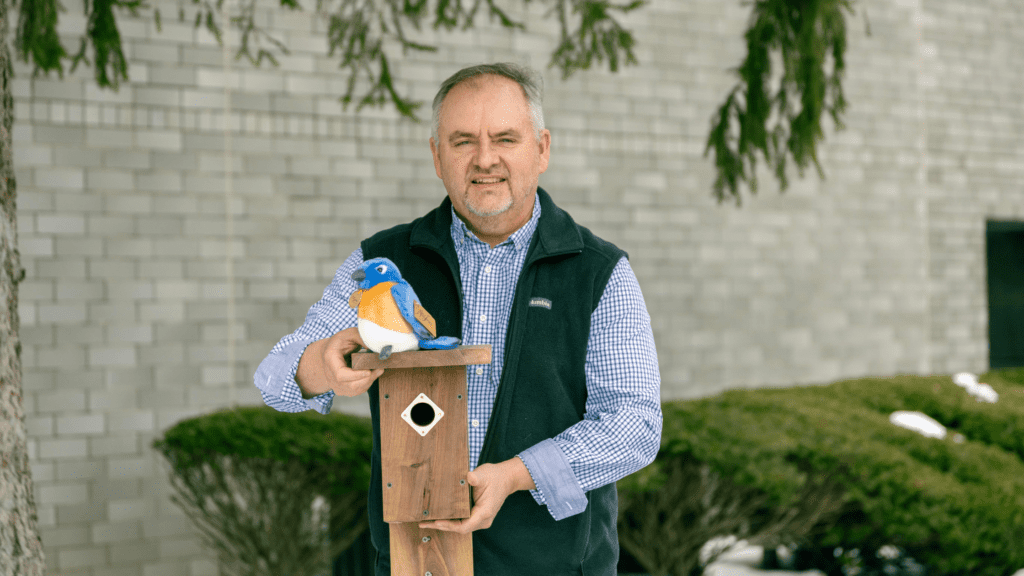 Man holding a wooden birdhouse with a plush bluebird on top, set against a brick wall and green shrubbery, showcasing bird conservation efforts.