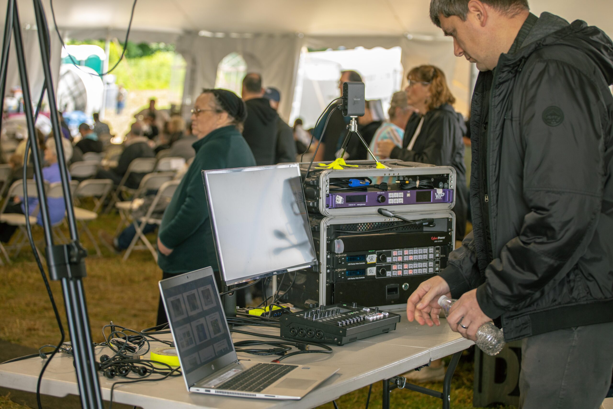 Technician operating audio-visual equipment at an outdoor event under a tent, with a crowd of attendees in the background, featuring monitors, a sound mixer, and various cables on a table.