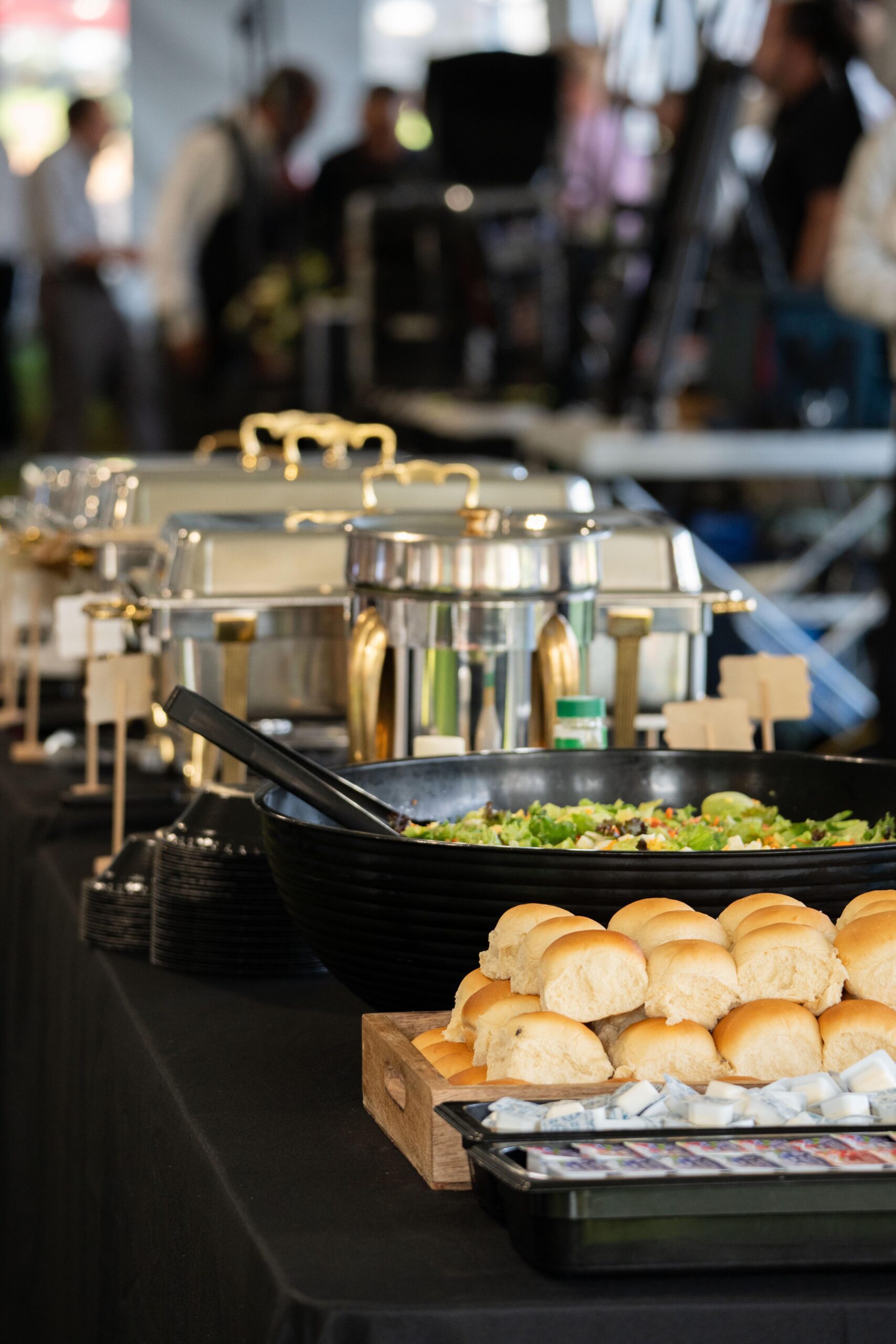 Catering buffet setup featuring a variety of dishes, including a large salad bowl, chafing dishes, and rolls arranged on a wooden tray, set against a blurred background of event attendees.