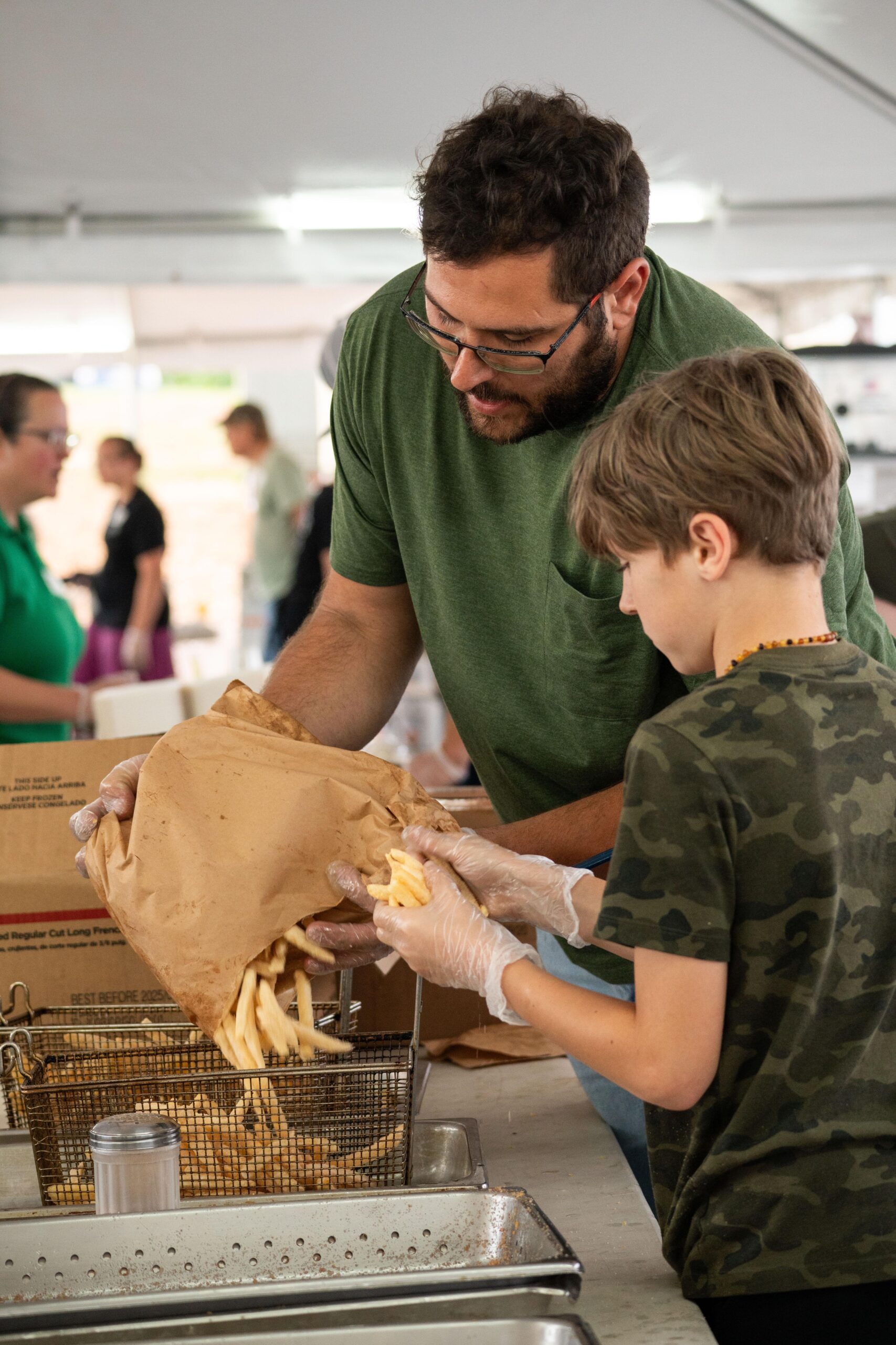 A man and a boy wearing gloves are preparing and serving French fries at a community event, showcasing teamwork and family involvement in food preparation.