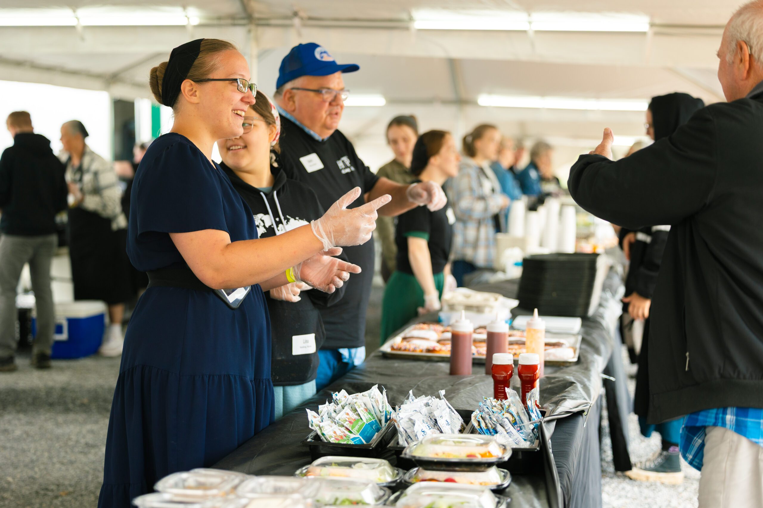 Community event featuring volunteers serving food at a tent. Smiling volunteers interact with attendees, showcasing various food items including packaged meals and condiments.
