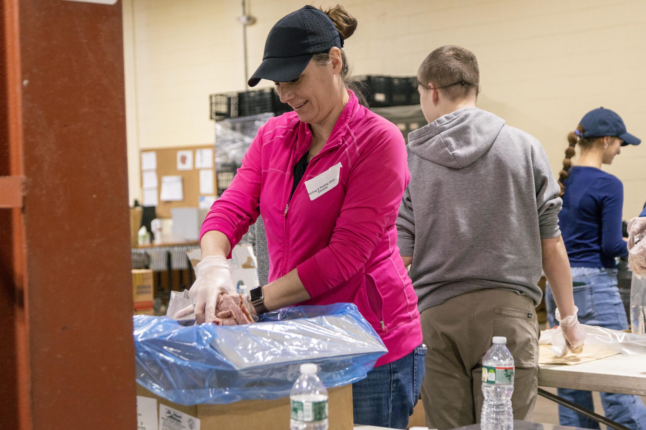 Volunteers packing food items at a community food bank, with a focus on a woman in a pink jacket using gloves to handle food, while others assist in the background.